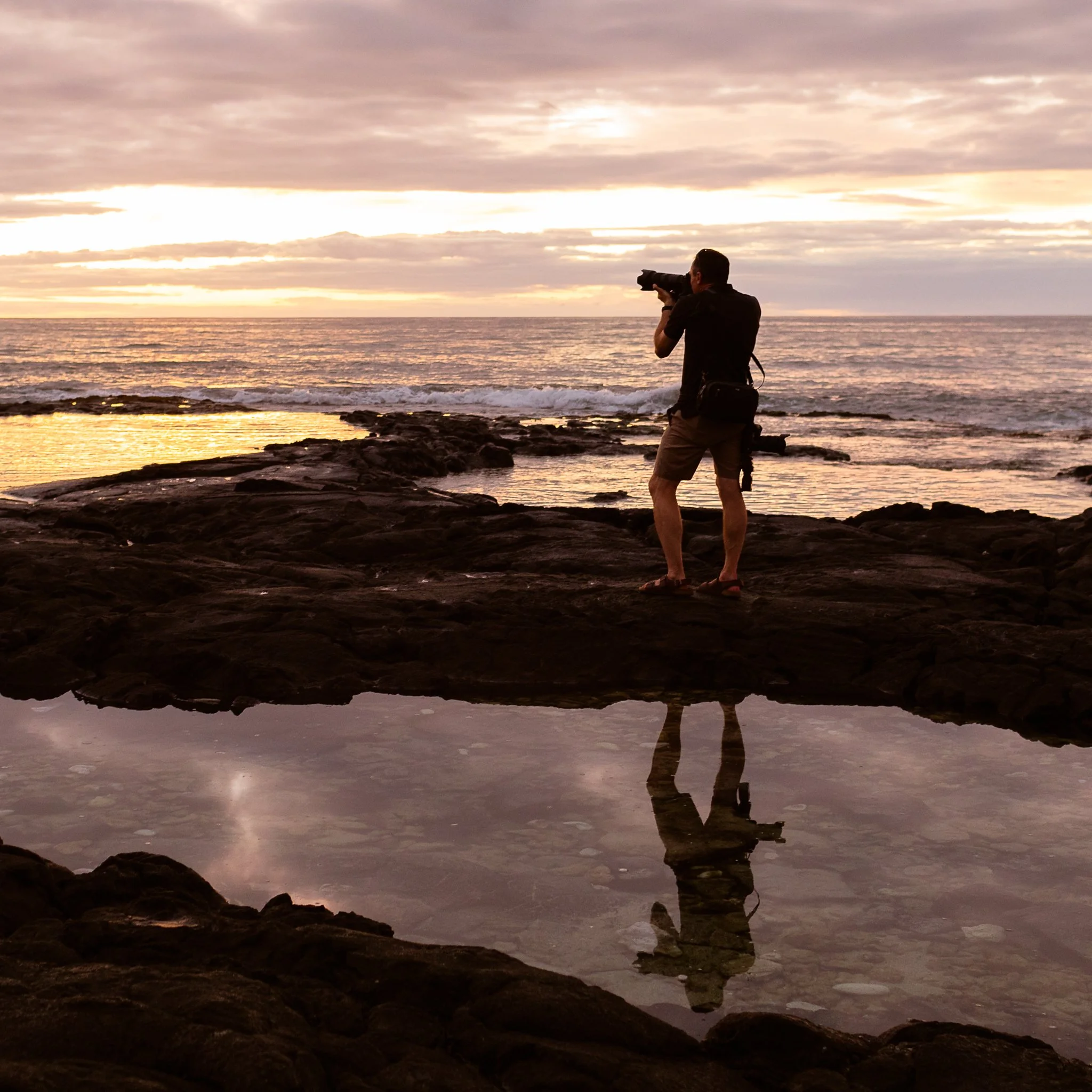 A person standing on rocks at the beach during sunset or sunrise, holding a camera and taking a photograph of the ocean and sky.