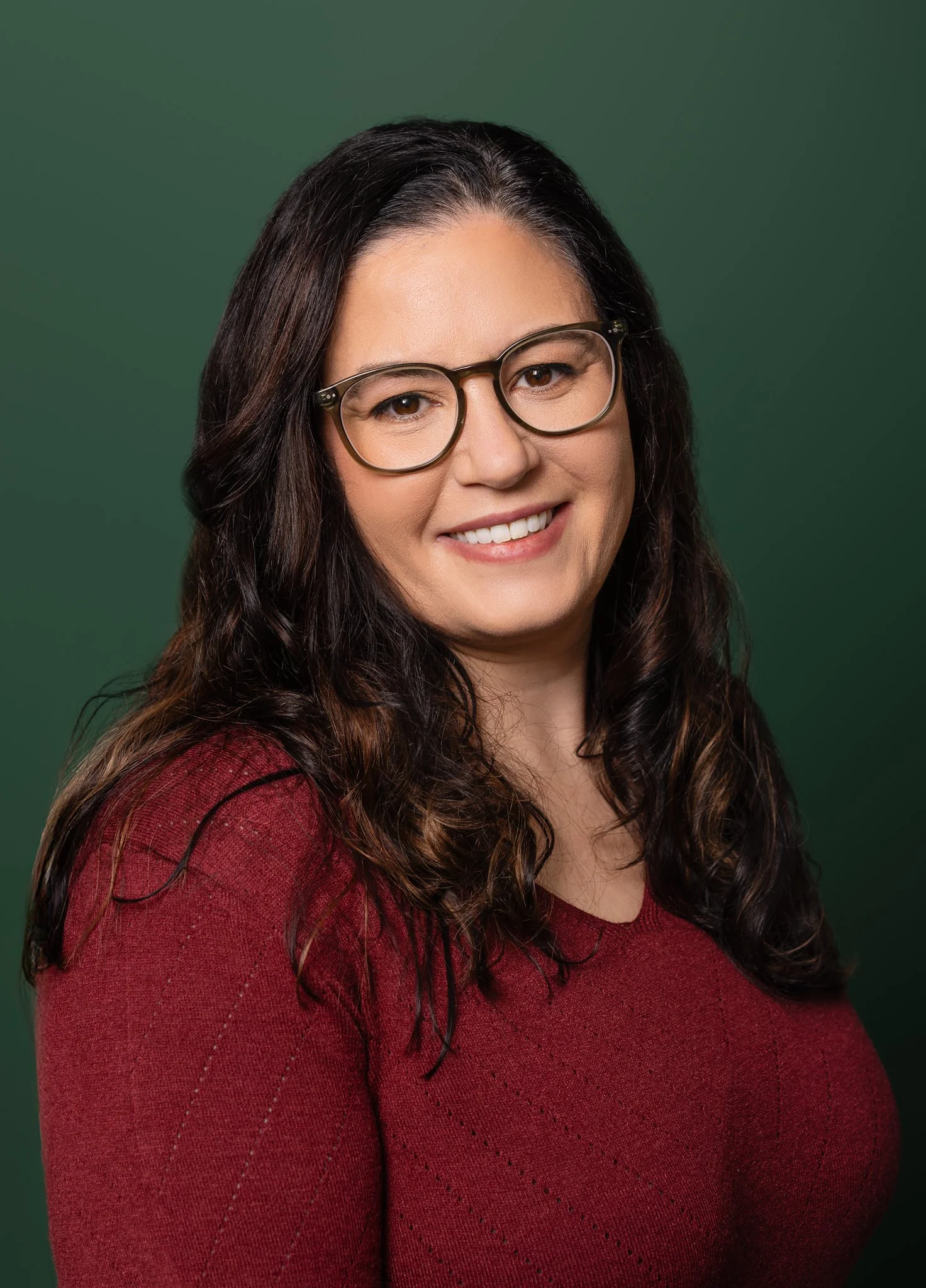 A woman with long dark hair wearing glasses and a red sweater, smiling against a green background.