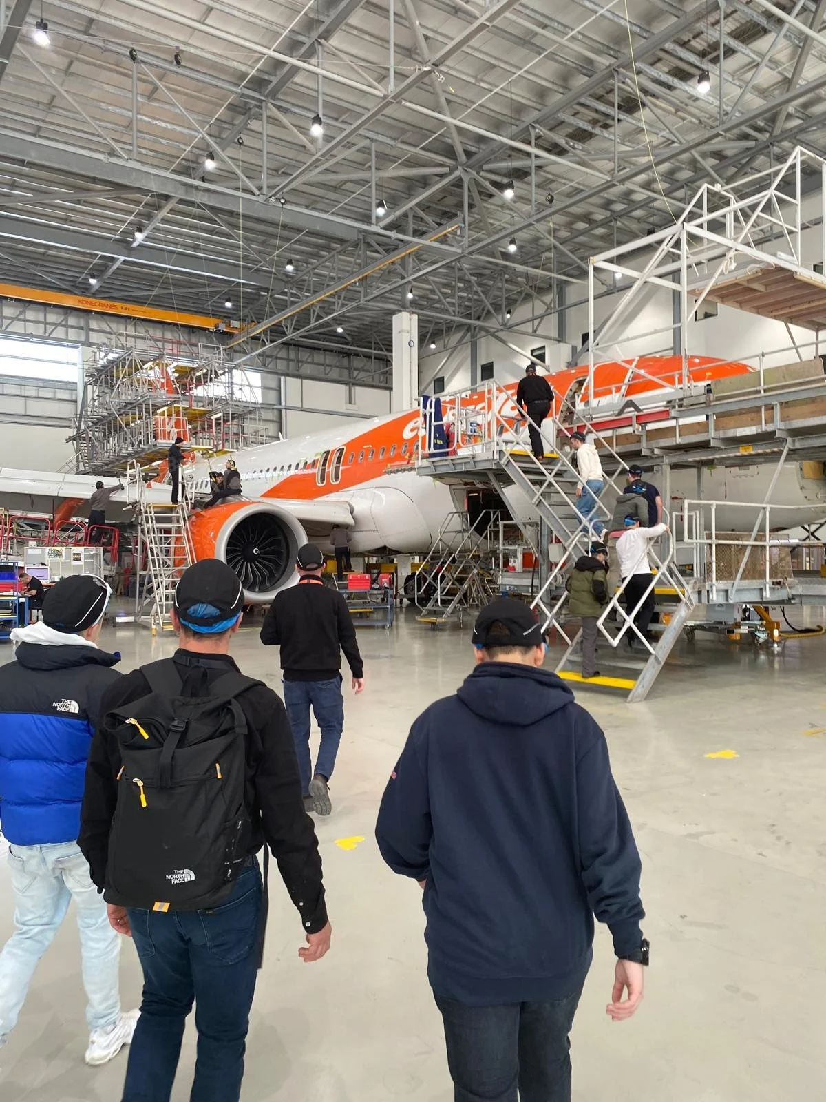 People walking inside an aircraft manufacturing or maintenance hangar, with a large airplane in the background being worked on by workers on elevated platforms.