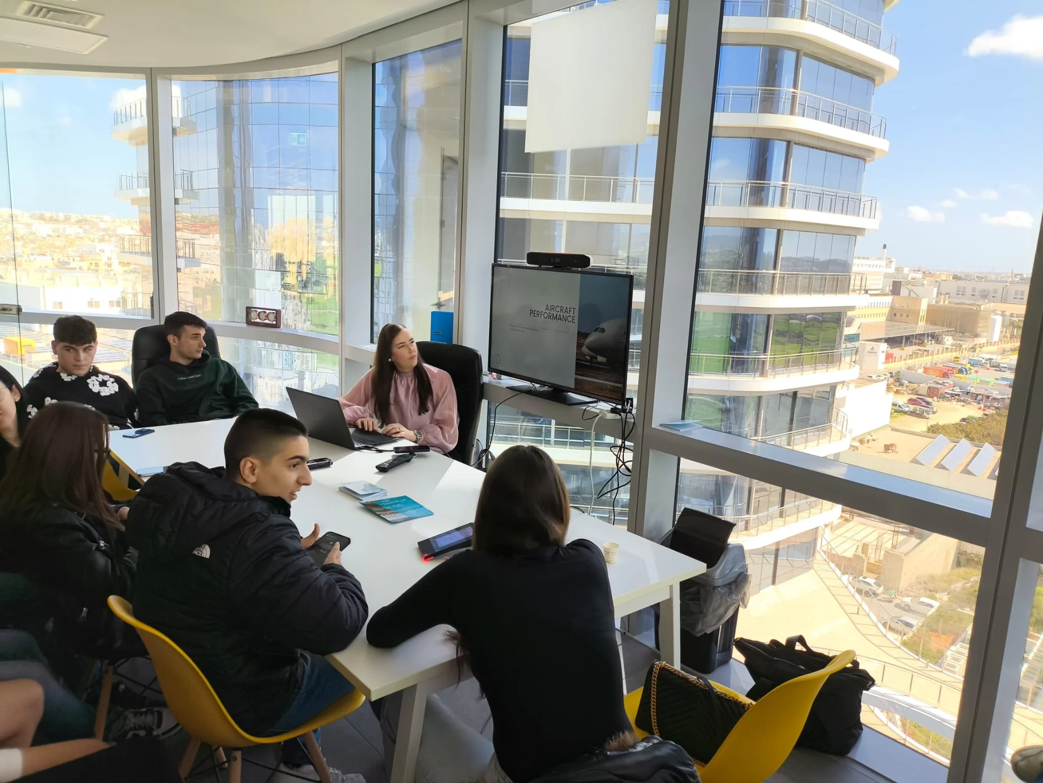 People sitting around a white conference table in a modern office, with large floor-to-ceiling windows showing a cityscape and a curved residential building outside.