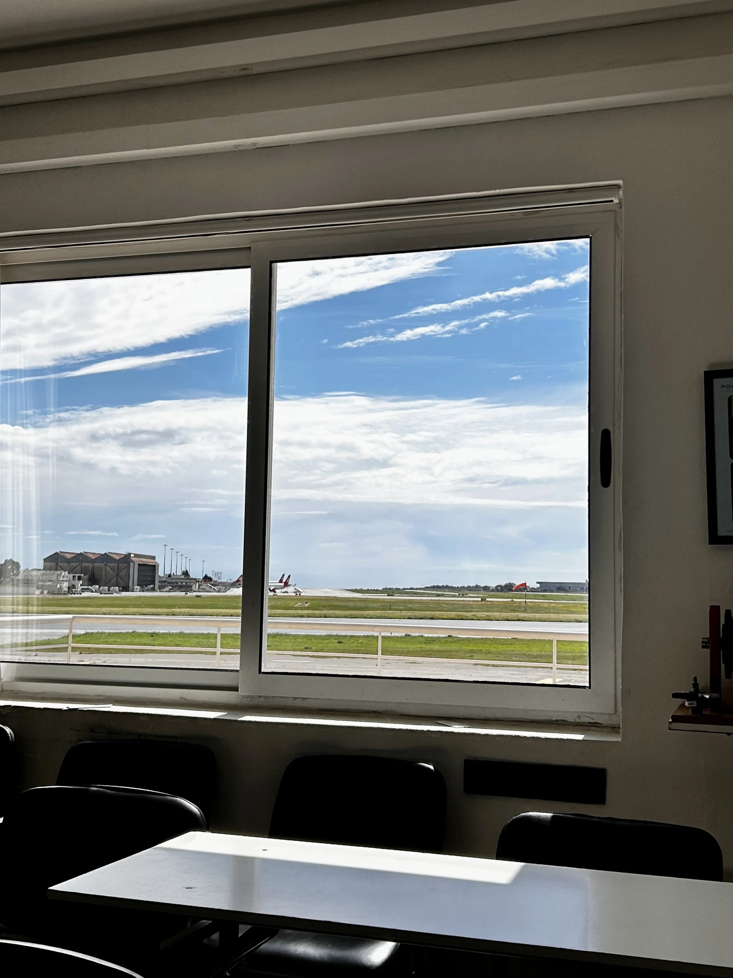 Airport terminal interior with large window view of runway, airplanes, and blue sky with clouds, taken from inside a room with chairs and a table.