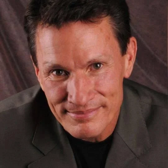 Close-up portrait of a man with dark hair, wearing a dark blazer, smiling softly with a wooden background.