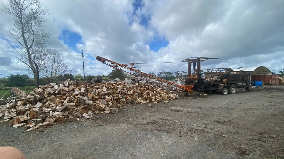 A wood processing yard with a large pile of chopped firewood, a wood splitter machine on a trailer, and a cloudy sky overhead.