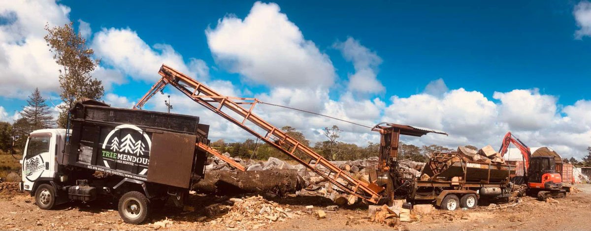 A logging truck and wood processing equipment at a wood yard, with a blue sky and white clouds overhead.