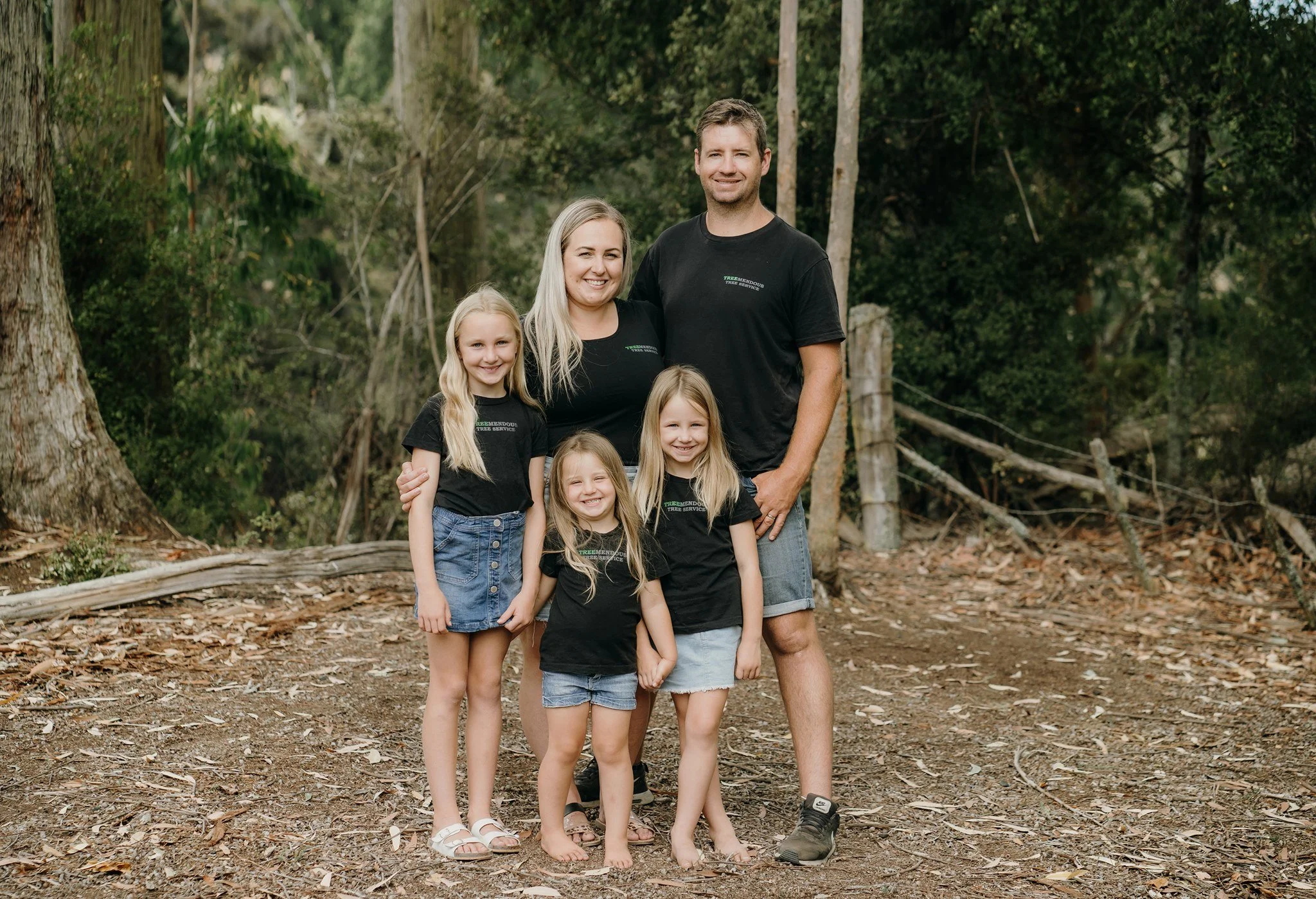 A family of six standing outdoors in a wooded area, smiling at the camera. The family includes two adults and four young girls, all wearing black T-shirts with a logo that reads 'TREEMENDOUS TREE SERVICE.' The background features trees and a forest floor covered with leaves and twigs.