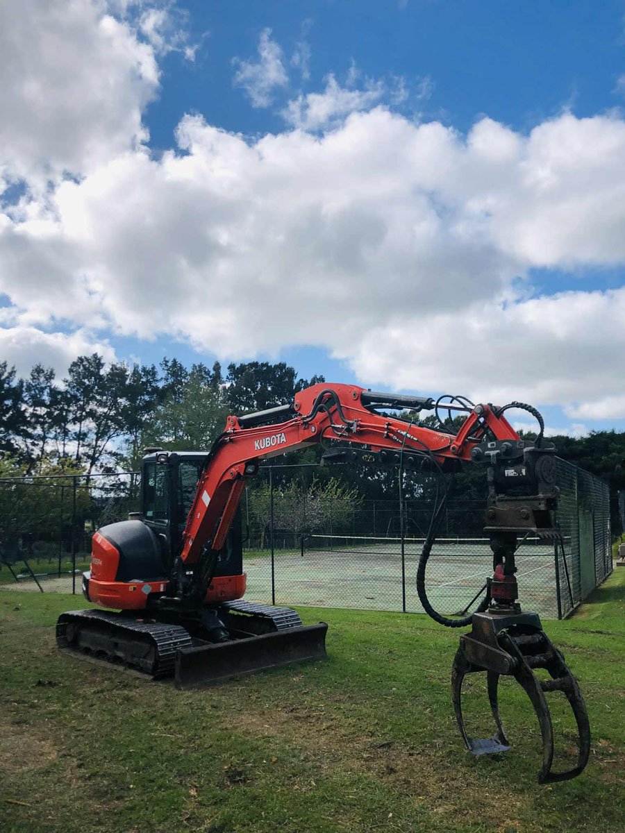 A small red Kubota excavator with a hydraulic arm and claw attachment parked on grass near a tennis court with a chain-link fence, trees, and a partly cloudy sky in the background.