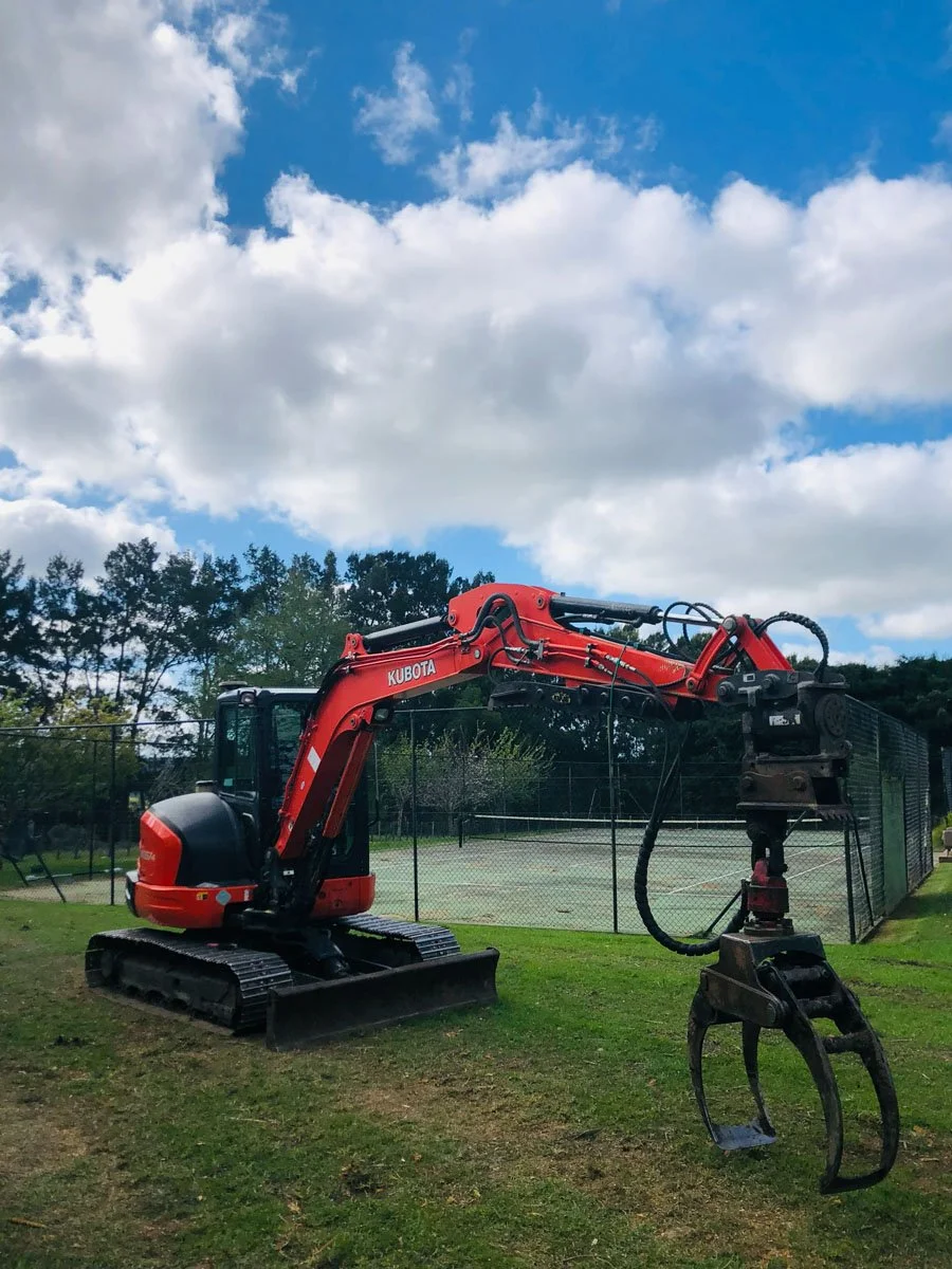 A red Kubota mini excavator with a grapple attachment parked on grass near a tennis court