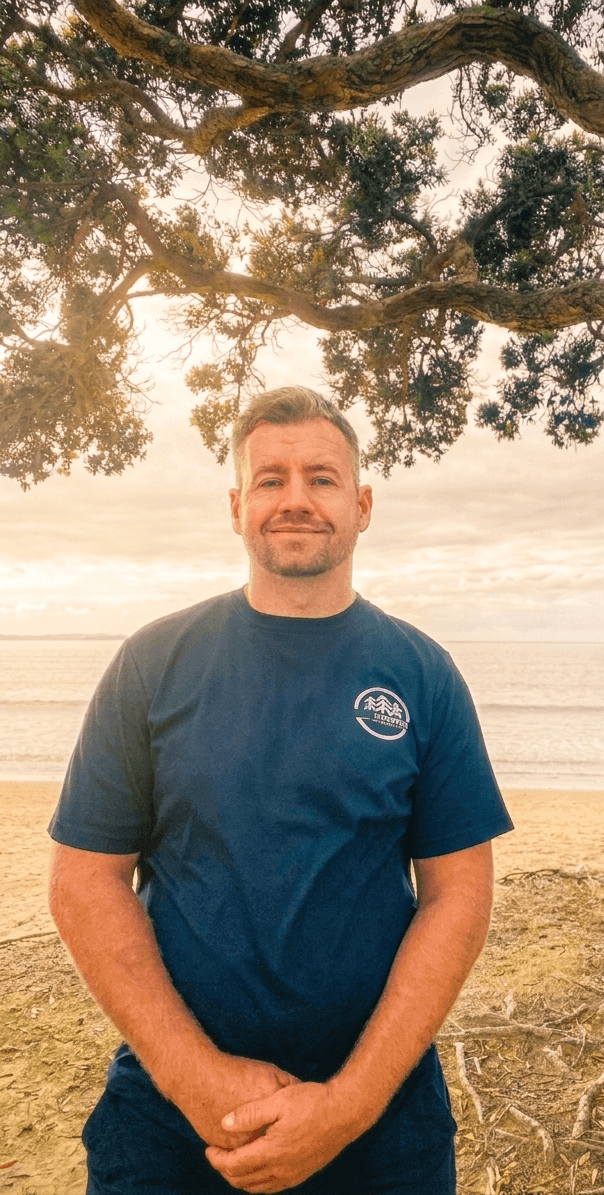 Man standing on a beach under a large tree at sunset, smiling, wearing a navy blue t-shirt with a mountain logo.