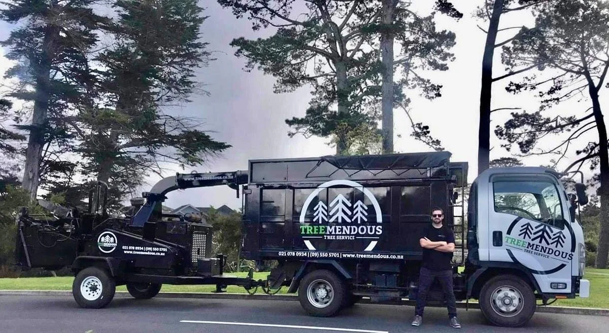 A man standing in front of a black tree service truck with the logo and contact information for Tree Mendous Tree Service parked on a suburban street with trees in the background.