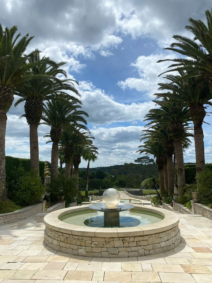A symmetrical stone fountain with a clear sphere sculpture on top, surrounded by palm trees, on a paved stone patio under a partly cloudy sky.