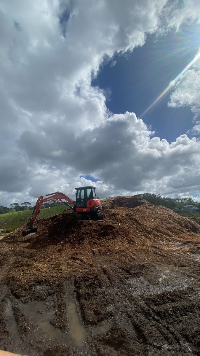 Construction site with a small orange excavator on a mound of dirt under a partly cloudy sky with sun rays.