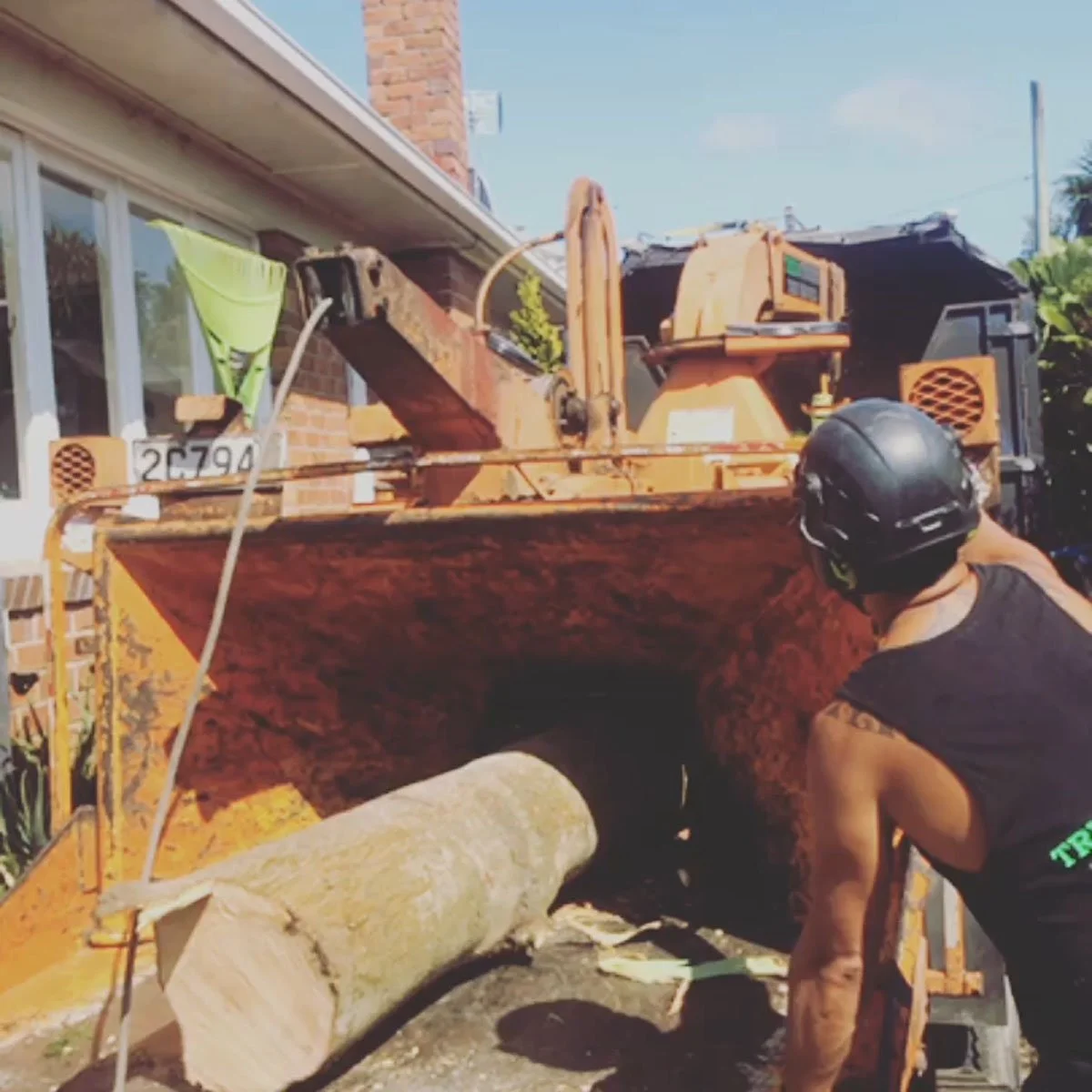 Worker wearing a helmet operating a wood chipper, feeding a large log into it outdoors near a brick house.