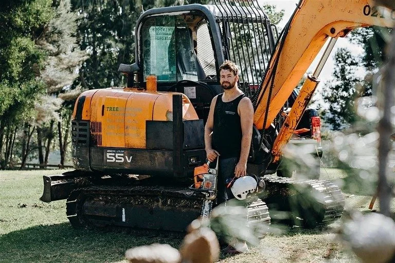 A man standing next to an orange and black mini excavator in a grassy outdoor area, holding a helmet and a chainsaw.
