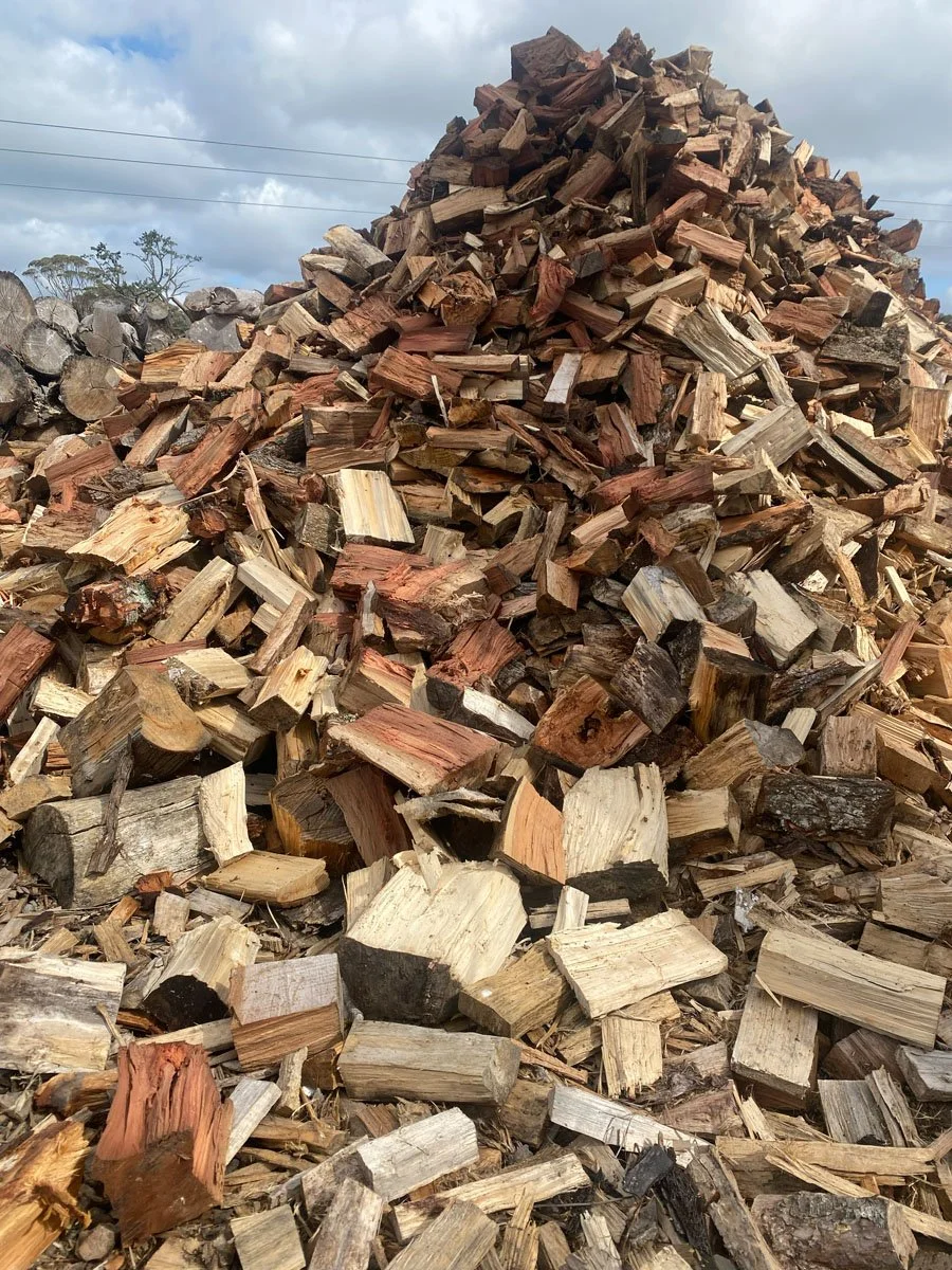 A large pile of chopped firewood with logs of various sizes, stacked outdoors against a cloudy sky.