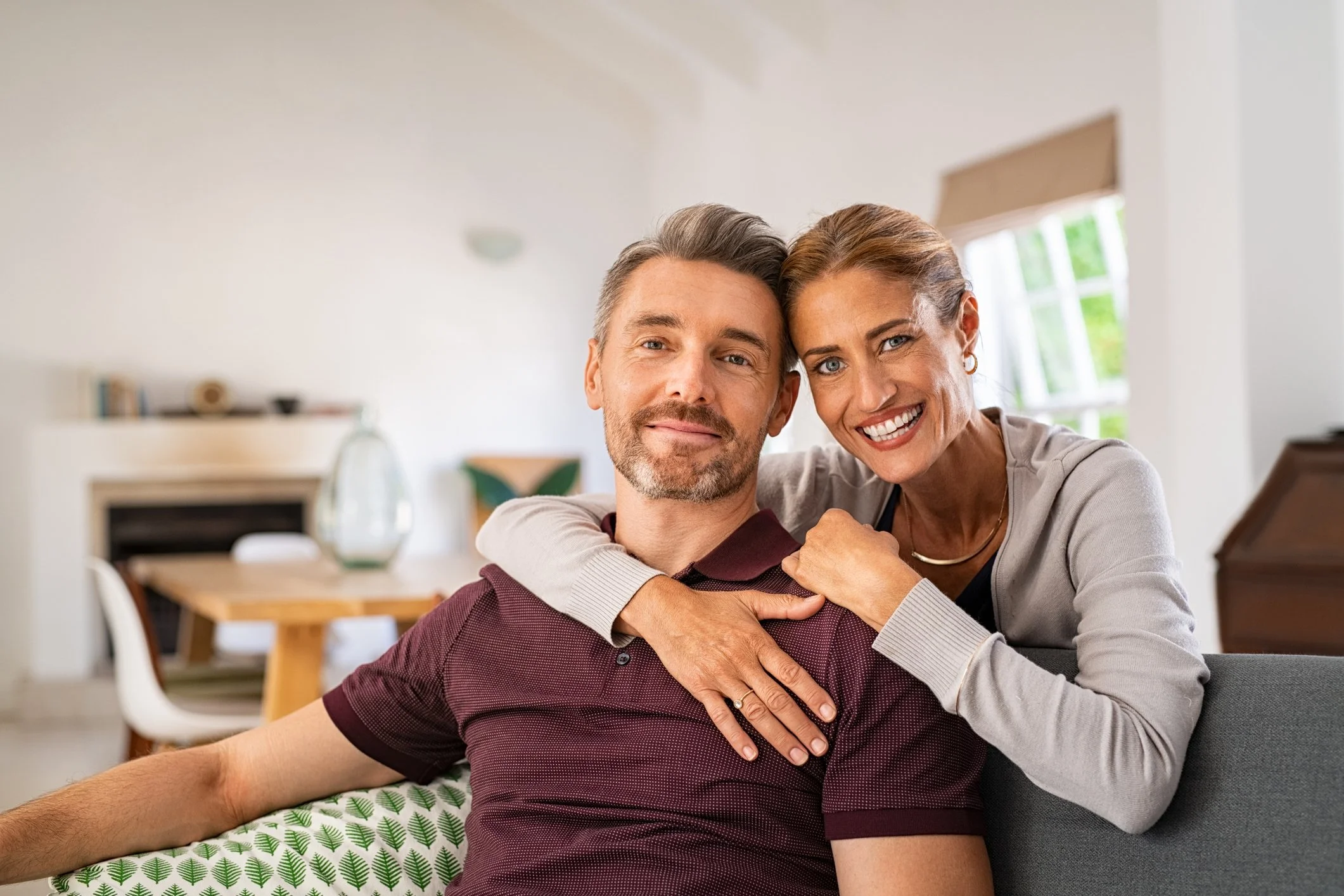 A middle-aged woman with short, light brown hair and a big smile is hugging and leaning her head on the forehead of a middle-aged man with salt-and-pepper hair and a beard, who is sitting and wearing a maroon shirt. They are in a bright room with large windows and a dining area in the background.