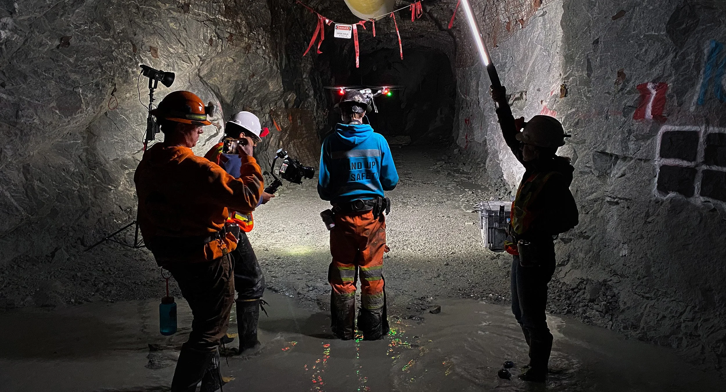Four people wearing helmets and safety gear inside a tunnel, with one person operating a drone, and the group standing in water near the tunnel floor.
