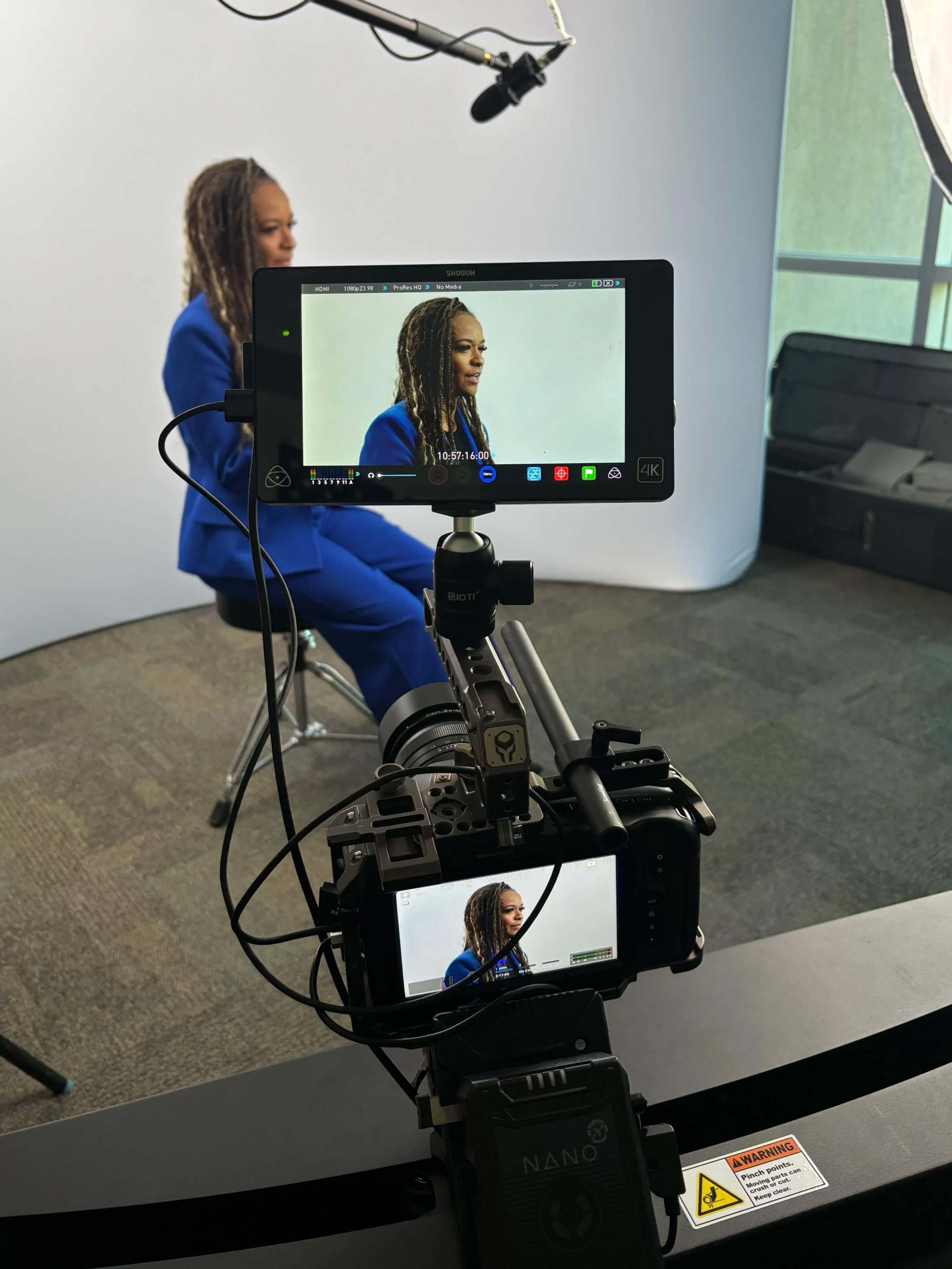 A woman in a blue suit sits on a stool in a studio with a white background, being filmed by a professional camera and monitor setup. The scene includes lighting equipment and a warning label on the table.