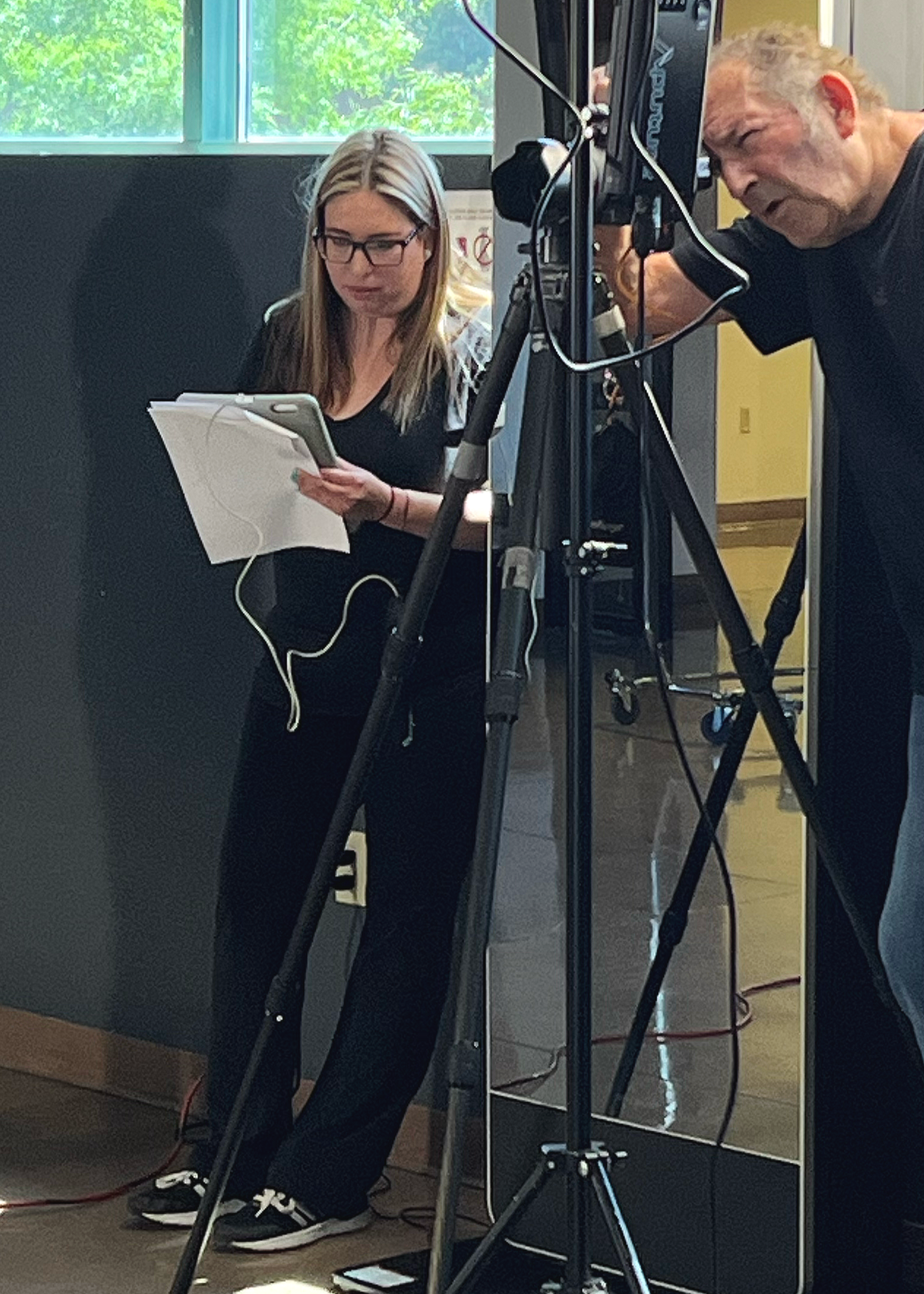A woman and an older man are working with professional video equipment in an indoor setting, with a large window showing green trees in the background.
