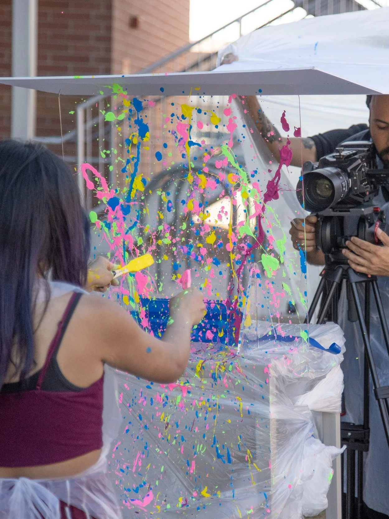 A woman is painting a colorful splatter design on a transparent sheet under a white canopy, with a man filming the scene using a professional camera.