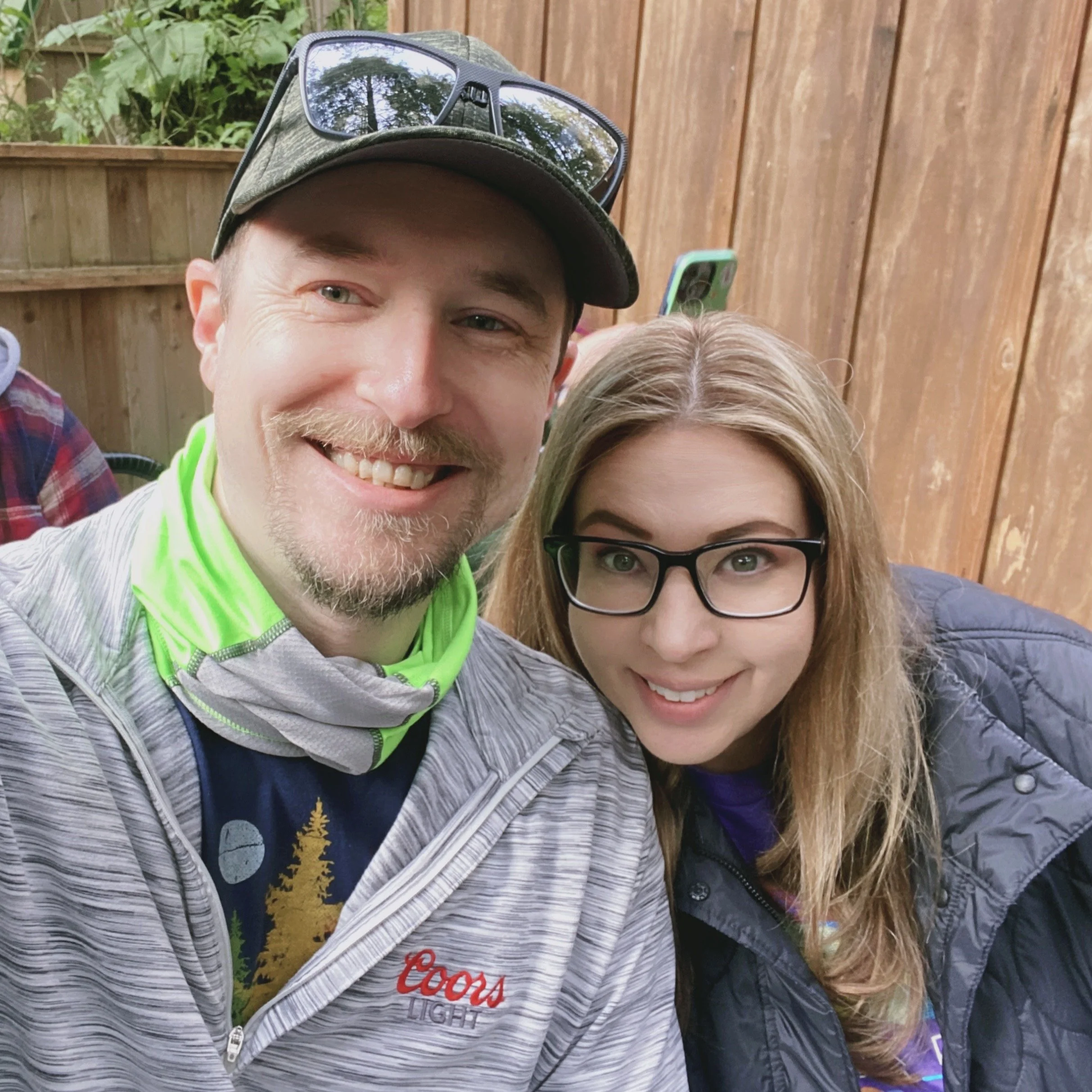 A smiling man and woman taking a selfie outdoors in front of a wooden fence, with glasses and casual clothing.