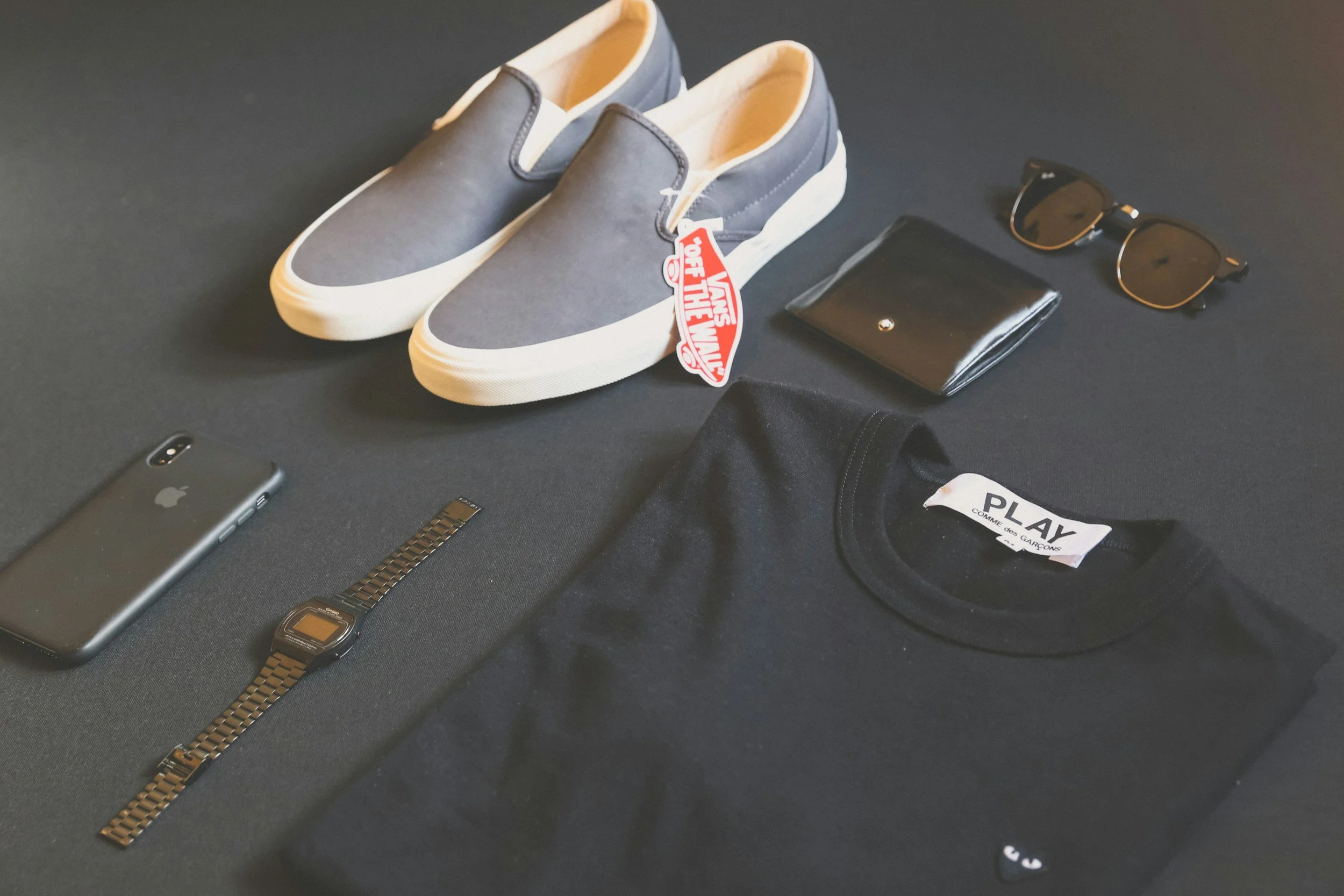 Black surface displaying various personal items including a pair of navy blue slip-on Vans shoes with a red Vans 'Out The Wall' tag, a black wallet, a pair of black and gold sunglasses, a black leather strap watch, a silver iPhone, and part of a black clothing item with a label that reads 'PLAY' and 'Comme des Garçons'.