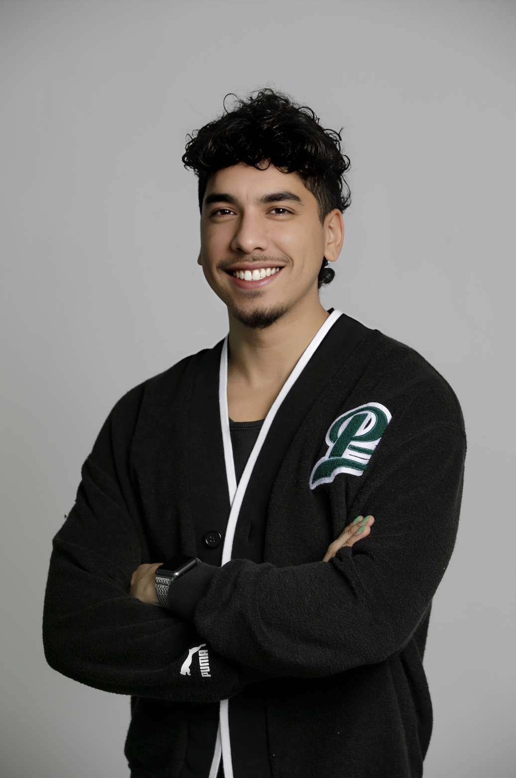 A young man with curly black hair and a goatee, smiling with arms crossed, wearing a black jacket with a patch and a smartwatch on his wrist, standing against a plain gray background.