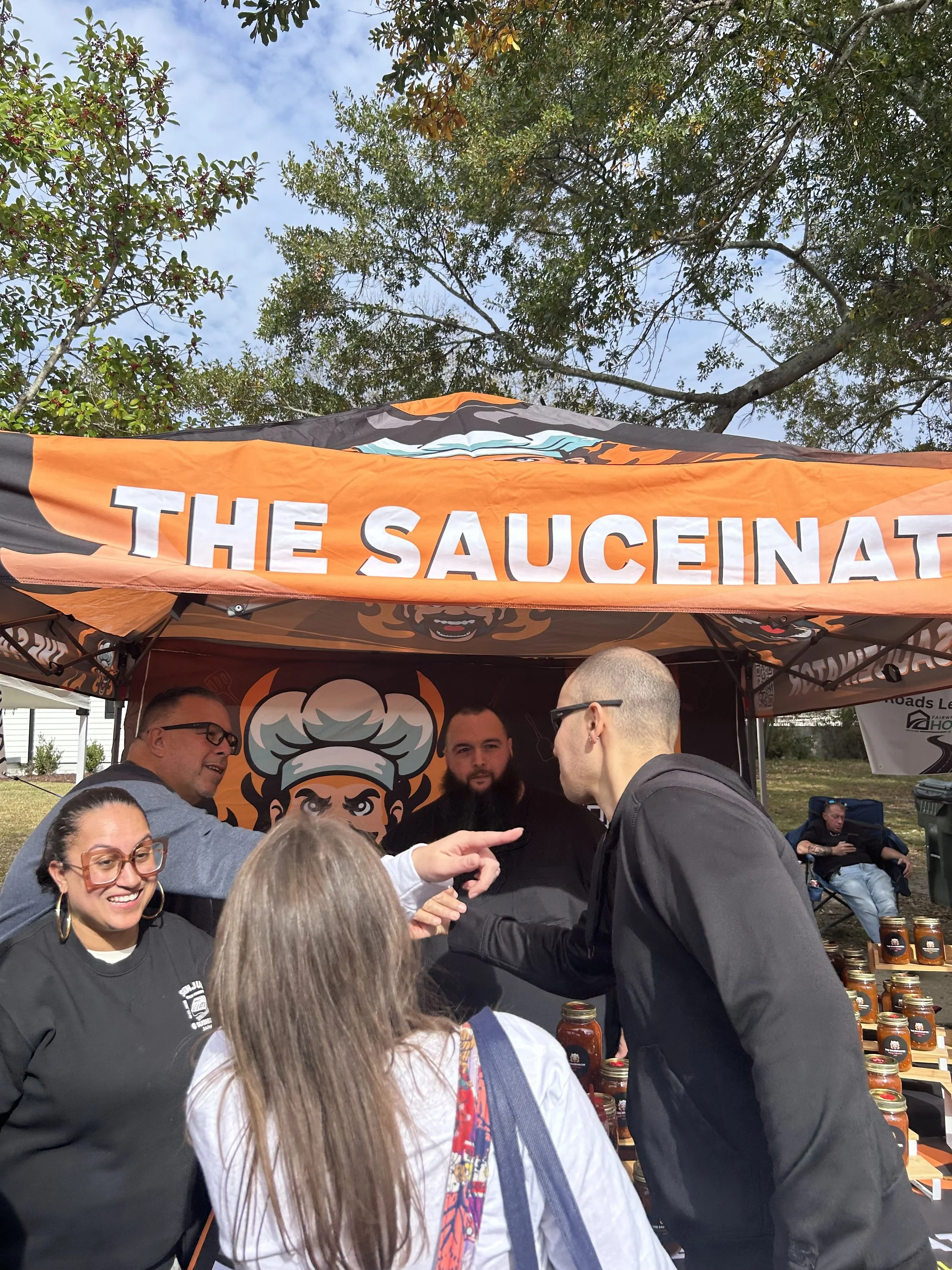 People gathered under a tent with a sign that reads "The Sauceinator," engaging in conversation at an outdoor event