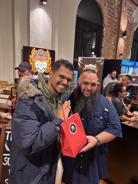 Two men smiling, standing close together inside a cafe or restaurant, with one holding a red gift bag. Other patrons and a neon lion logo are visible in the background.
