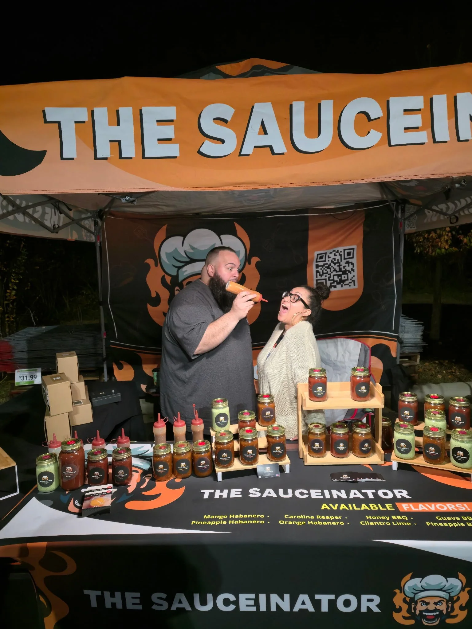 A man and woman at a stall called 'The Sauceinator' at night, with jars of sauce on display, and the man is tasting sauce from a squeeze bottle, while the woman smiles.