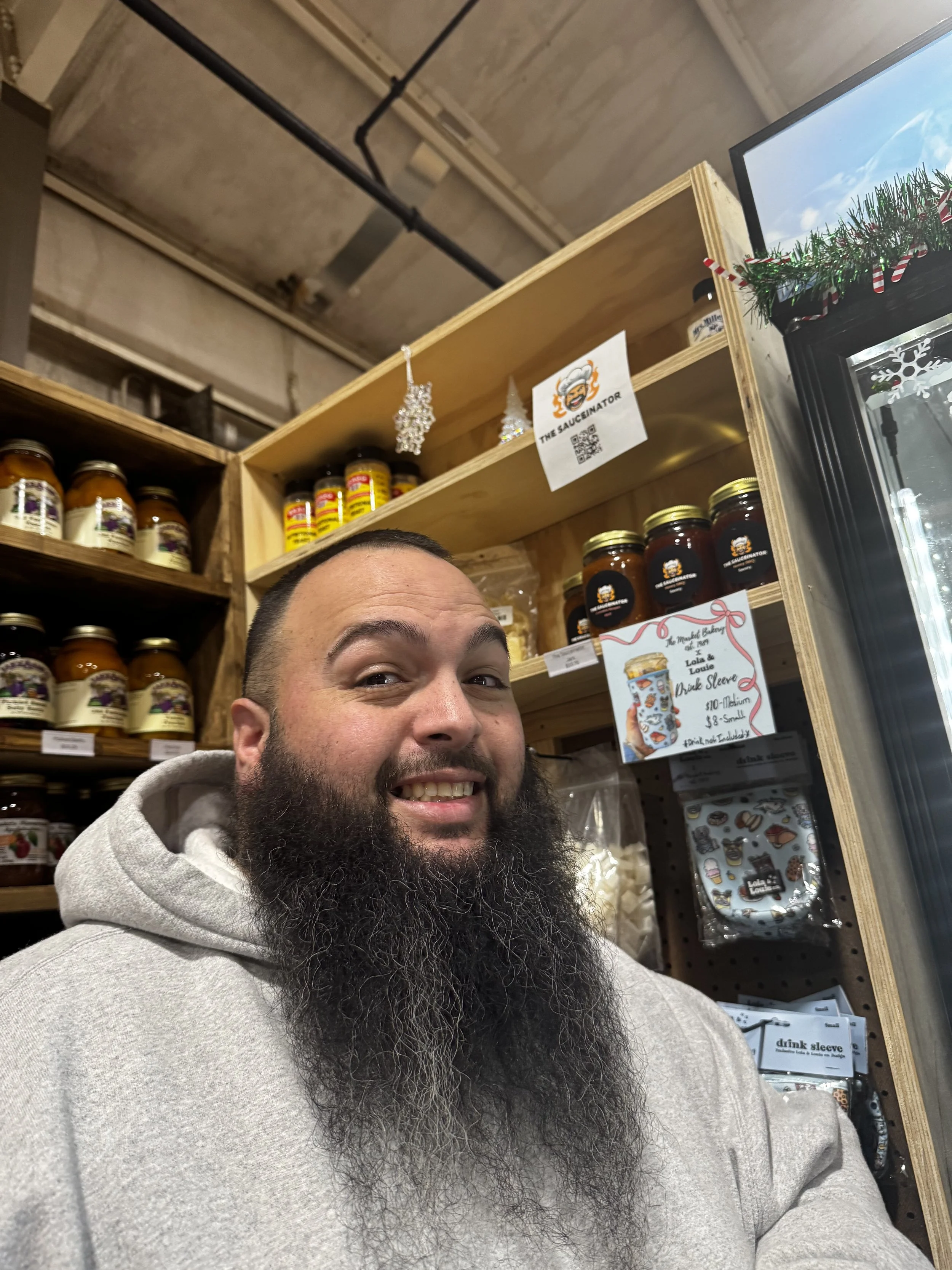 A man with a long beard smiling in front of shelves filled with jars and containers, with a refrigerator and holiday decorations visible in the background.