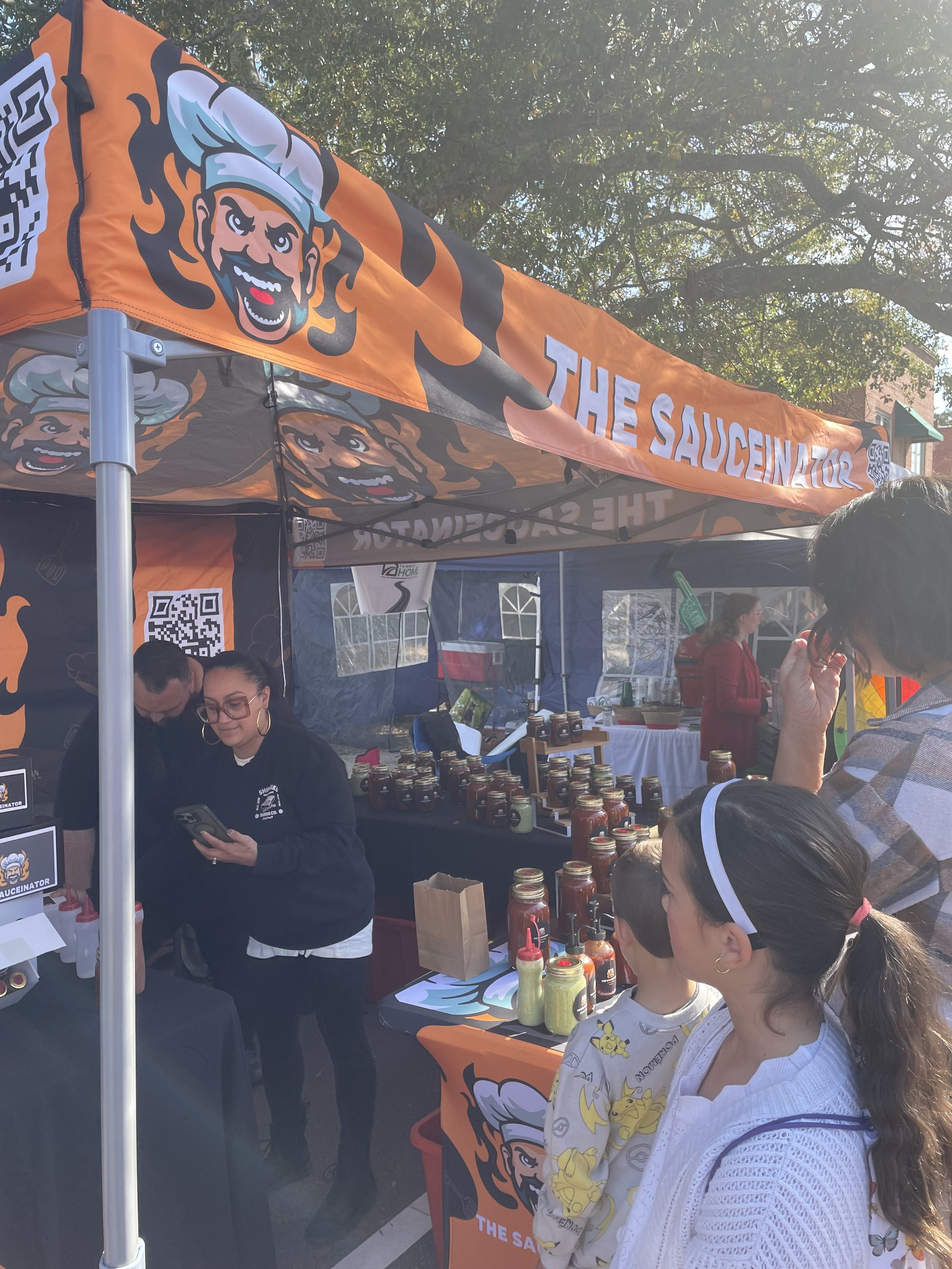 A food stall called "The Saucenator" with an orange canopy featuring a cartoon chef logo. People are gathered around, looking at jars of sauces on display, with two children and several adults present.