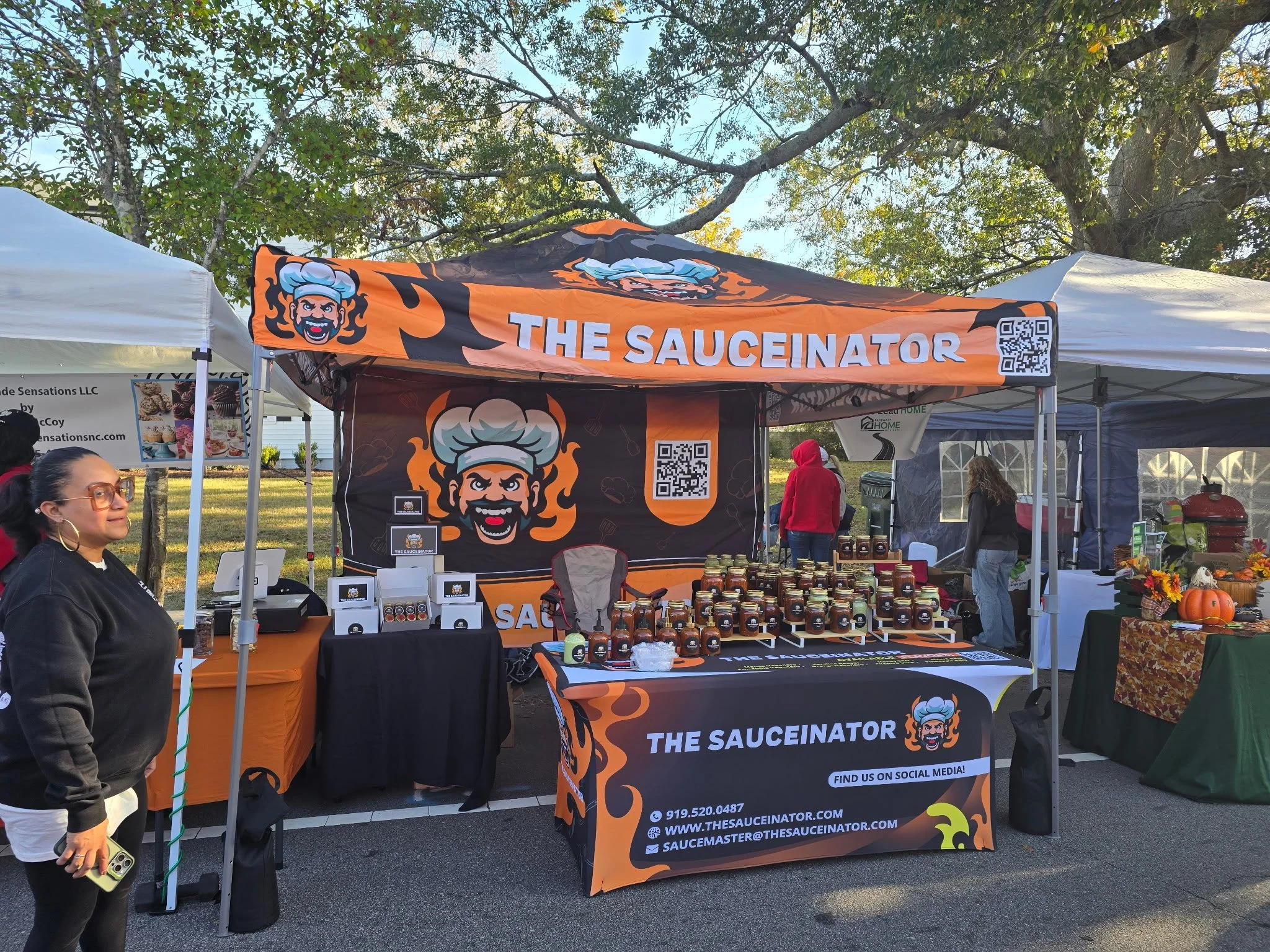 Outdoor market booth for 'The Saucinador' with a banner and table displaying hot sauces, surrounded by other vendor tents and people, with trees and a cloudy sky in the background.