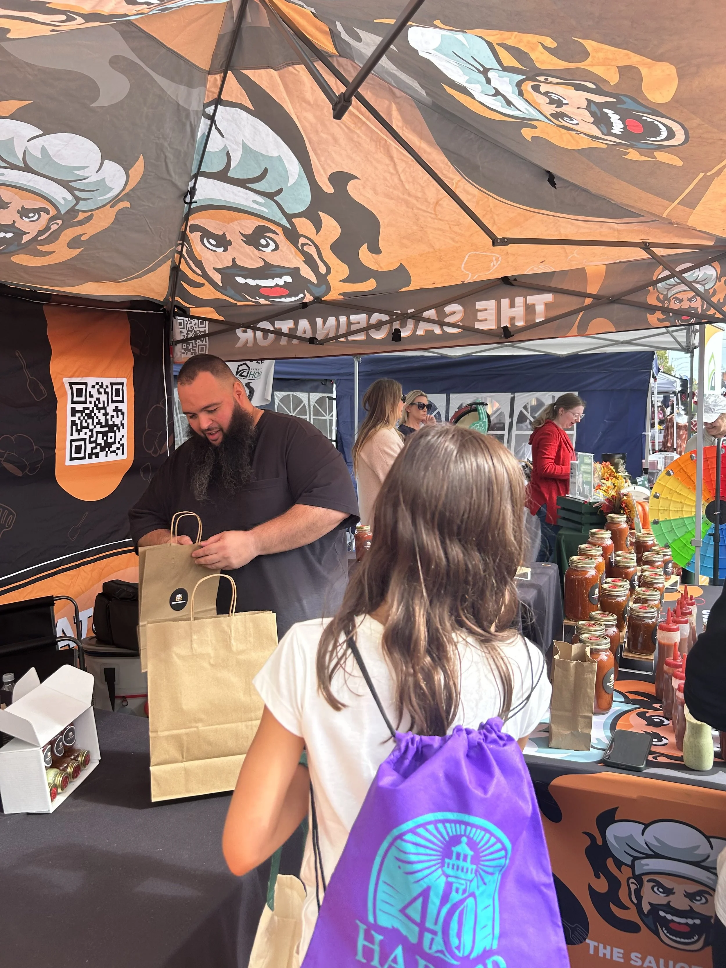 A vendor at an outdoor market stand serving customers, with jars of sauce on display and a picnic-style tent overhead.