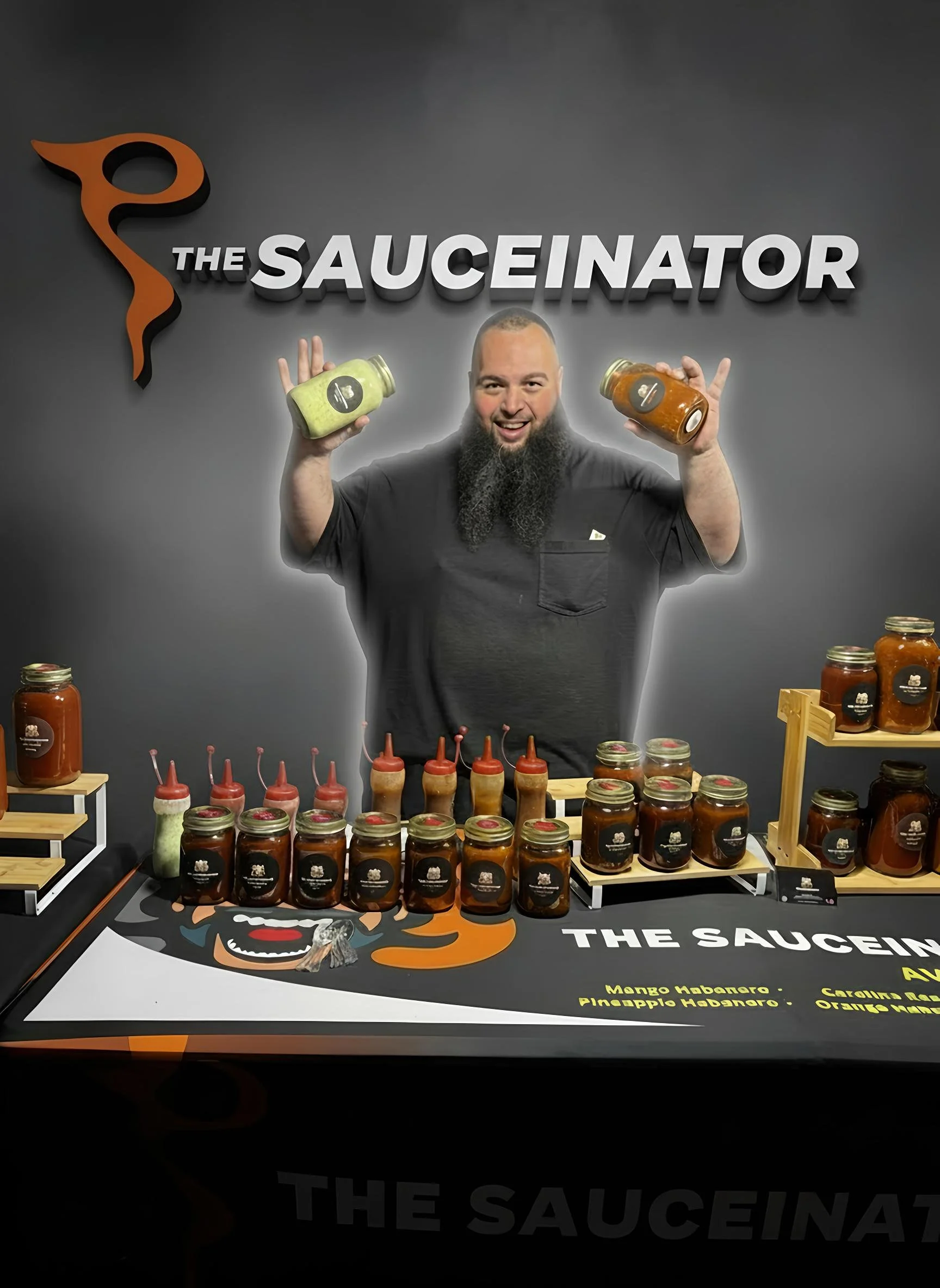 Man with beard displaying jars of sauce, standing behind a table with various jars and bottles of sauce, with a logo featuring a chili pepper and the text "The SauCeinator" on a gray background.