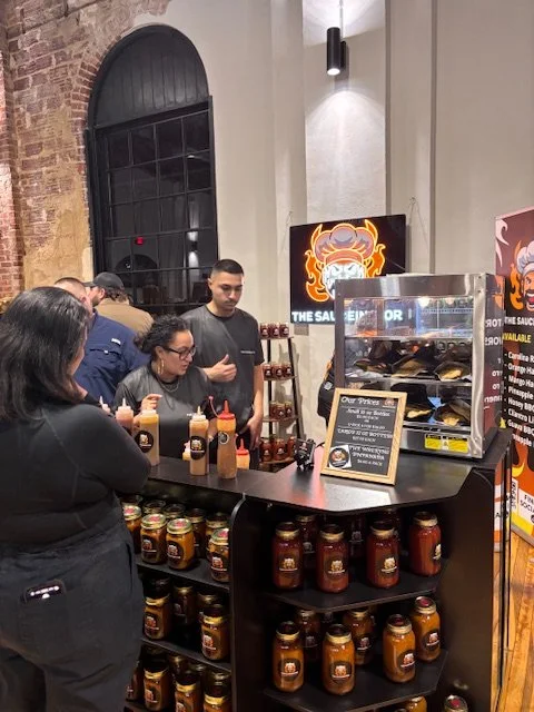 People in line at a food stand with a display case, sauces, and jars of preserved food in a modern industrial-style setting with a dragon logo on the wall.