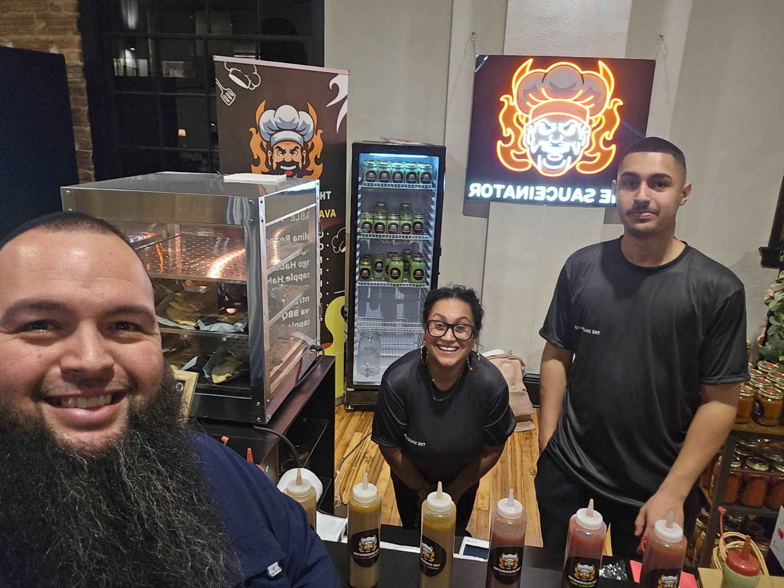 Group of three people behind a counter at a food stand, with bottles of sauces, a food warmer with food, and a refrigerator in the background, inside a restaurant or food stall.
