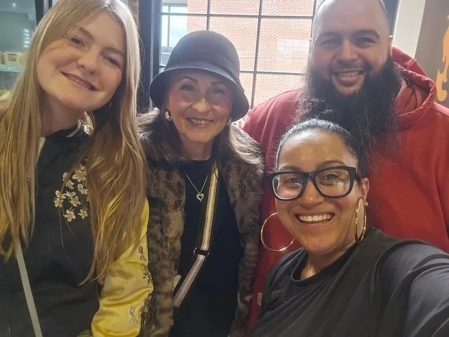 Four people smiling for a selfie indoors, two women and two men, with large windows and an urban brick building in the background.
