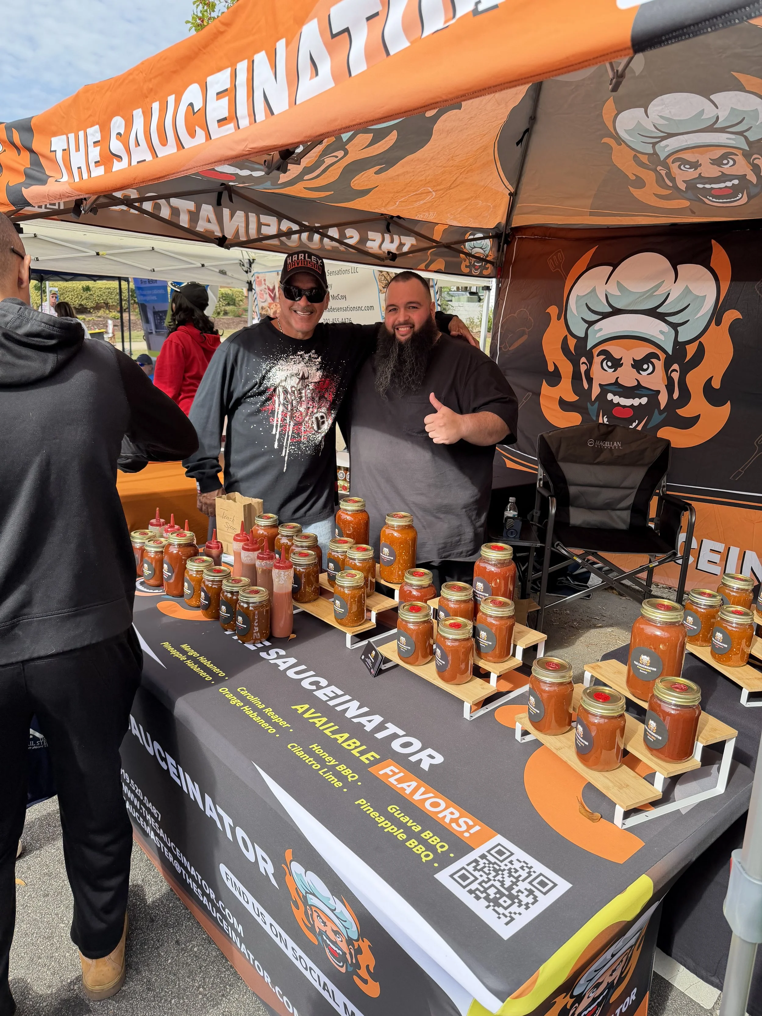Two men standing behind a table at an outdoor booth for Sauceinator with jars of barbecue sauce on display, smiling. One man is giving a thumbs-up. The booth has an orange and black canopy and a large logo of a chef's face with flames.