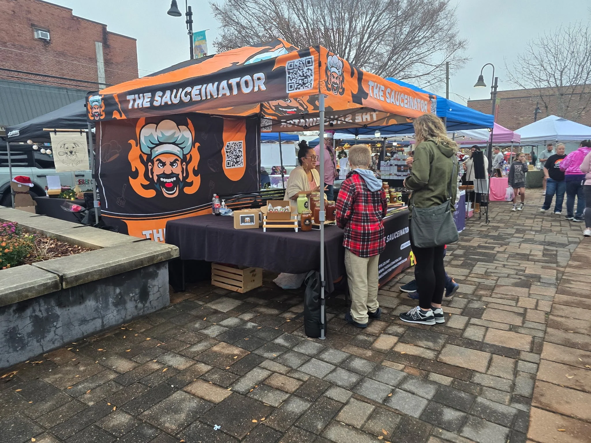 People shopping at an outdoor market booth called 'The Saucinator' with a cartoon chef logo, orange and black banner, and jars of sauce on display.