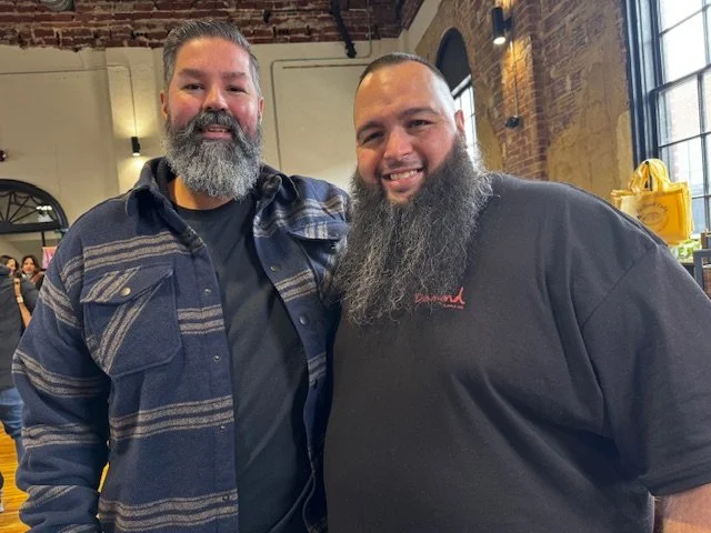 Two men with beards smiling indoors, one wearing a checkered jacket and the other in a black t-shirt, in a rustic brick-walled space with large windows.