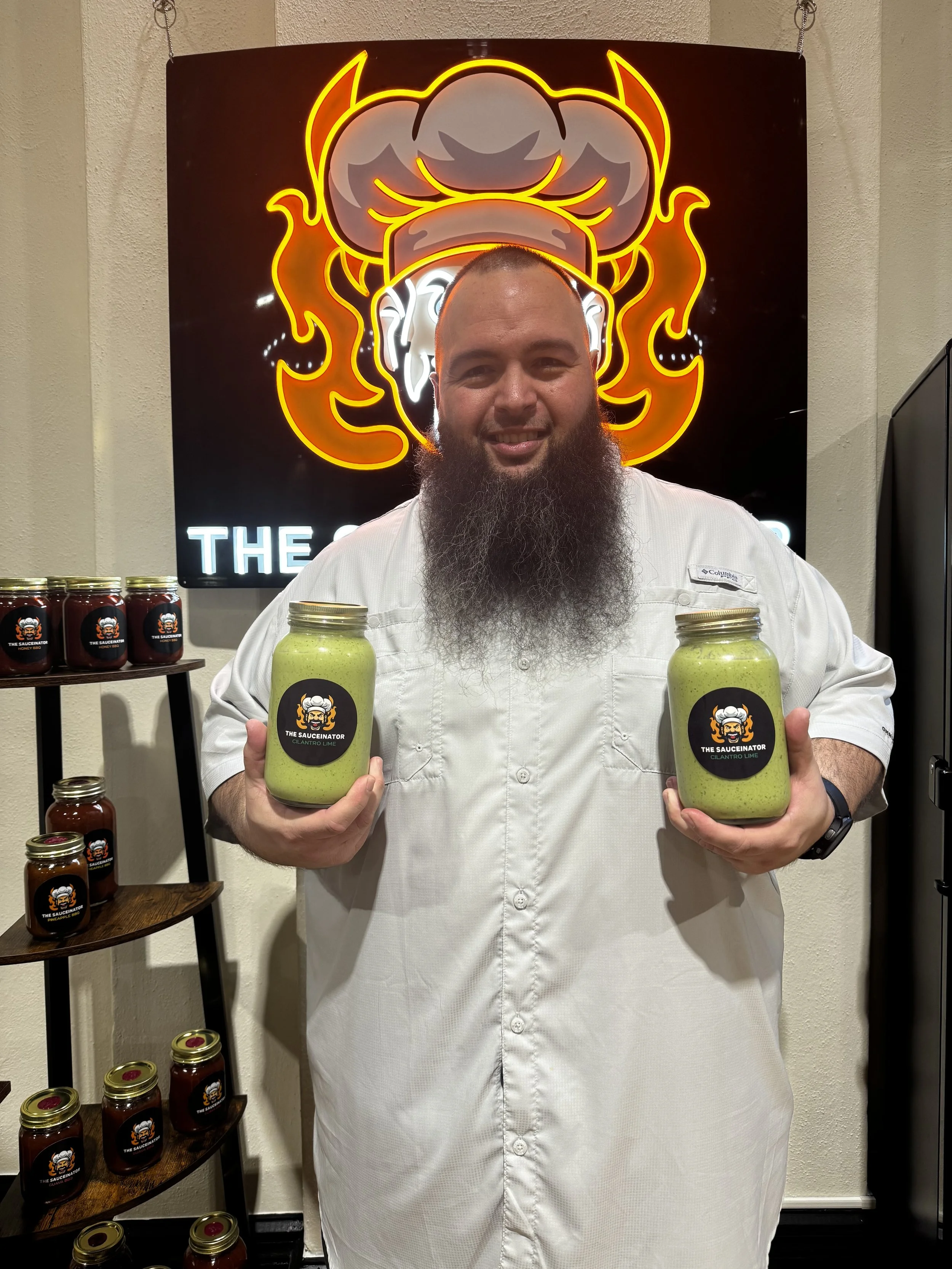 Man with a long beard holding jars of green sauce labeled 'The Sauceinator Cilantro Lime' standing in front of a neon sign with a chef's hat and flames.