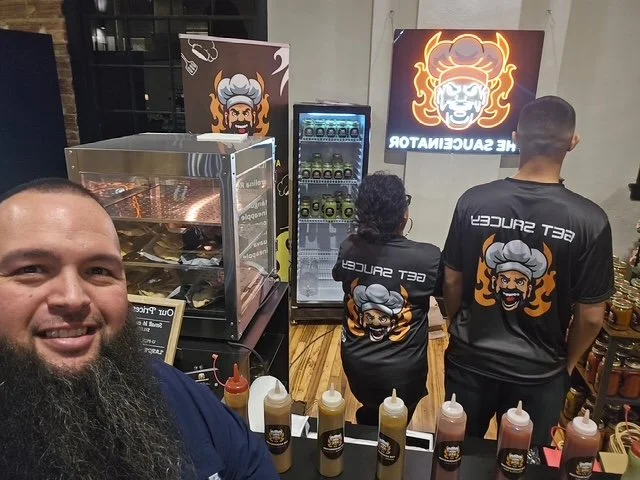 Inside a restaurant or food stall, three staff members wear black shirts with a cartoon chef logo and the words 'Get Sucked' visible. A man takes a selfie, capturing a popcorn machine, condiments, a beverage refrigerator, and a illuminated neon sign 
