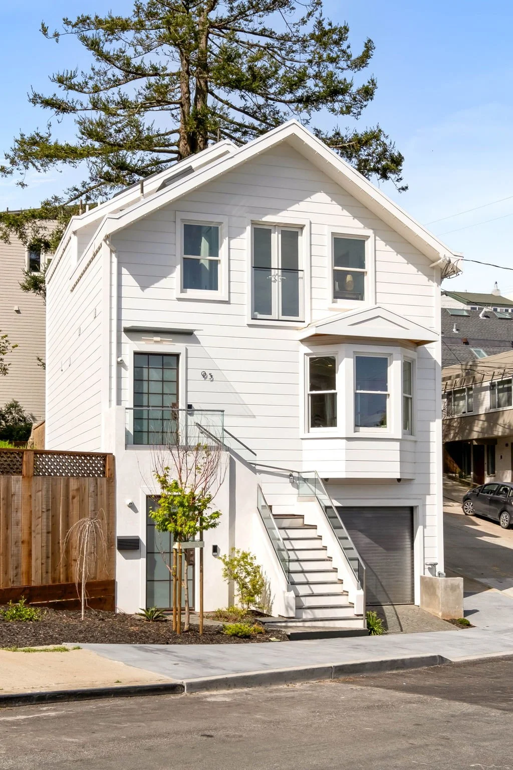 A three-story white house with a staircase outside leading to a front door. The house has multiple windows, a garage at the bottom, and a large tree behind it. A wooden fence and a small landscaped area are in the front.