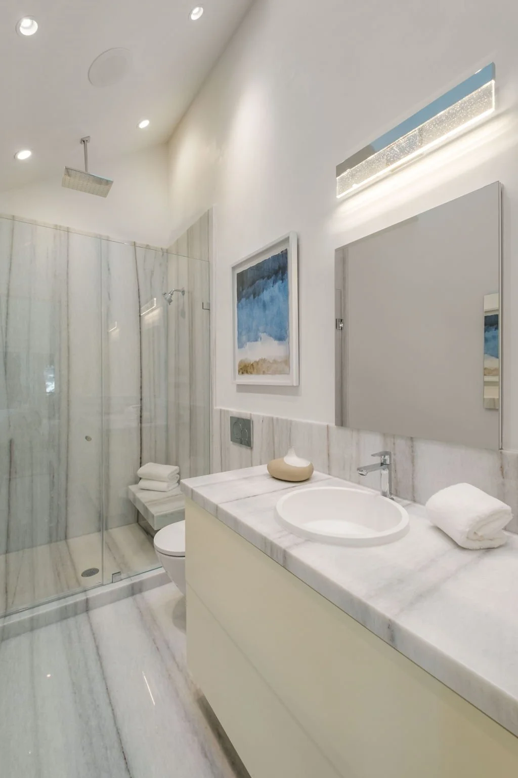 Modern bathroom with a marble countertop, round sink, and a large mirror. There is a shower with glass doors, wall art, and folded towels.