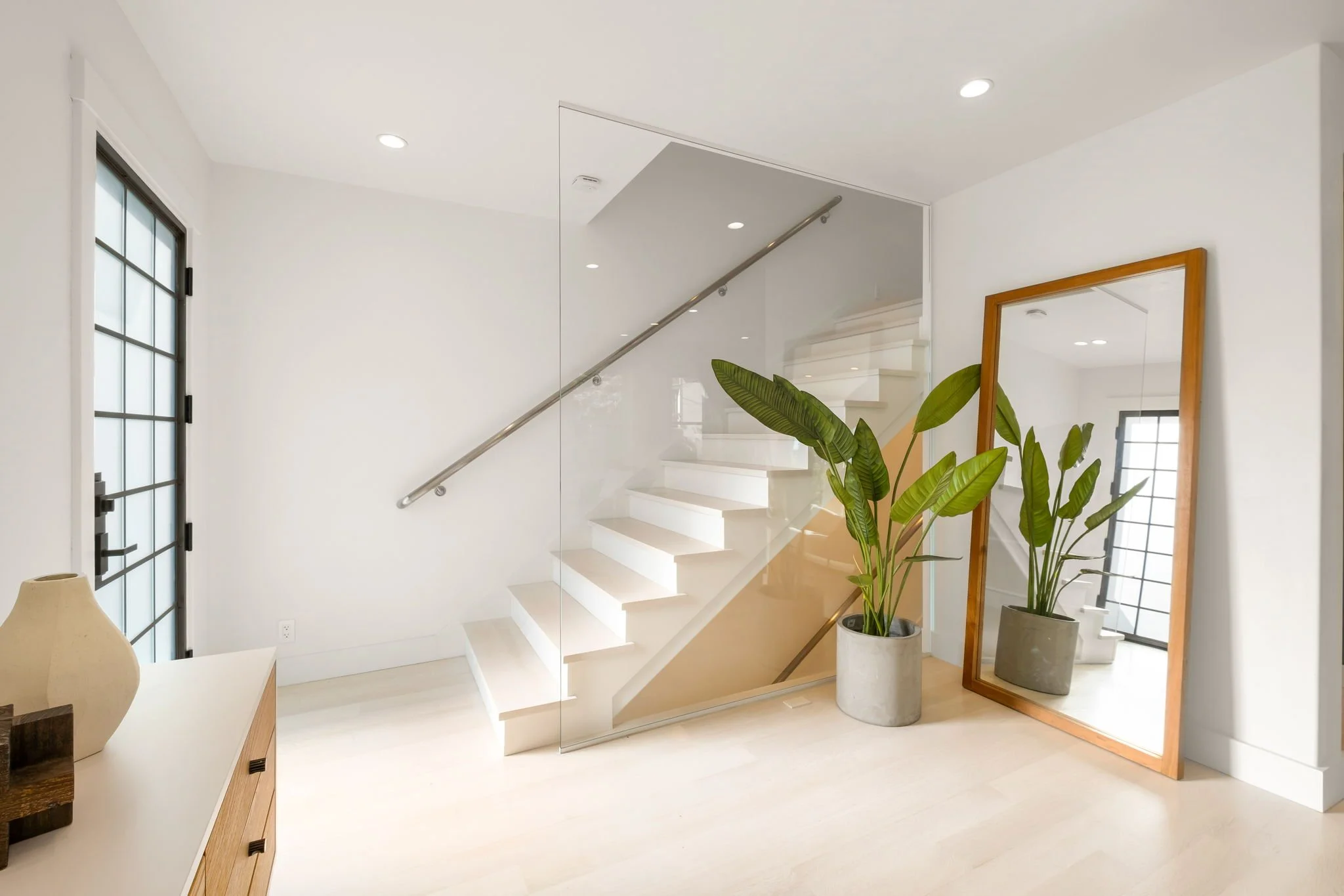 Minimalist interior with white walls, a staircase with glass railing, a large mirror with a wooden frame, a potted plant, and a door with a grid design.