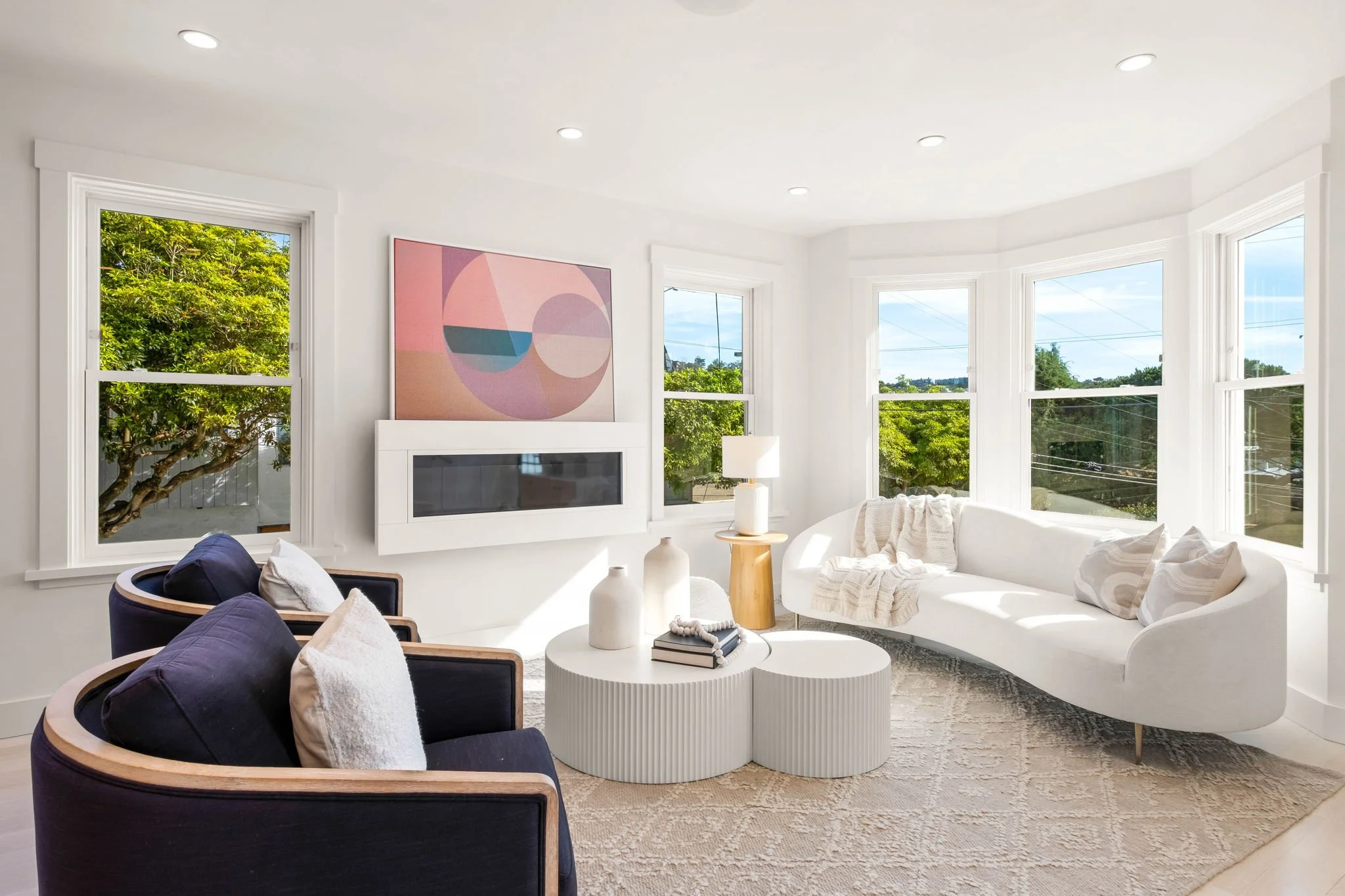 Bright living room with white walls and large windows showing green trees outside, featuring a curved white sofa, two dark armchairs, a round coffee table with vases and books, a side table with a lamp, and a pastel abstract painting on the wall.