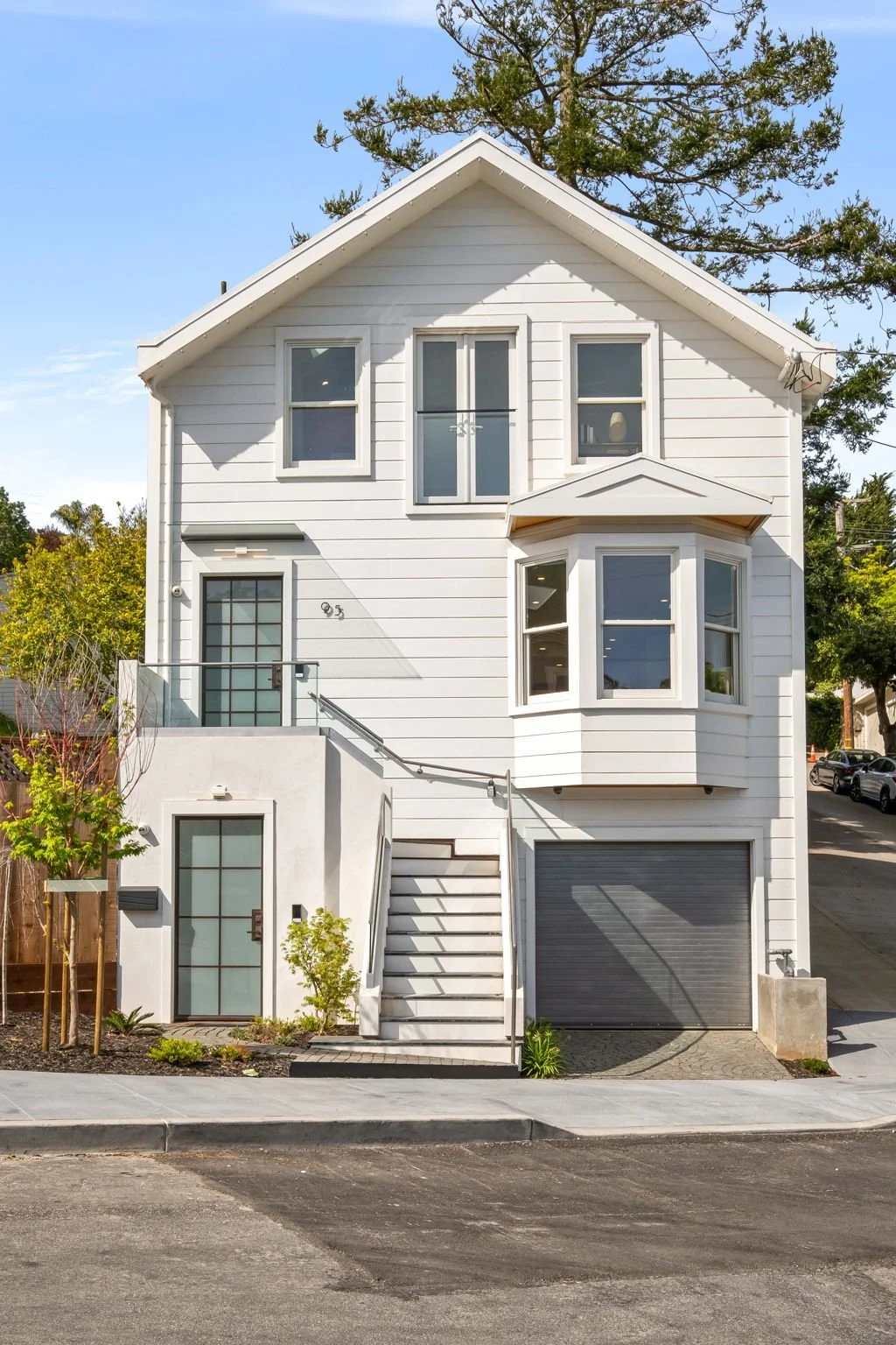 A modern three-story white house with a garage, stairs leading to a glass door, and multiple windows, surrounded by a small landscaped front yard.