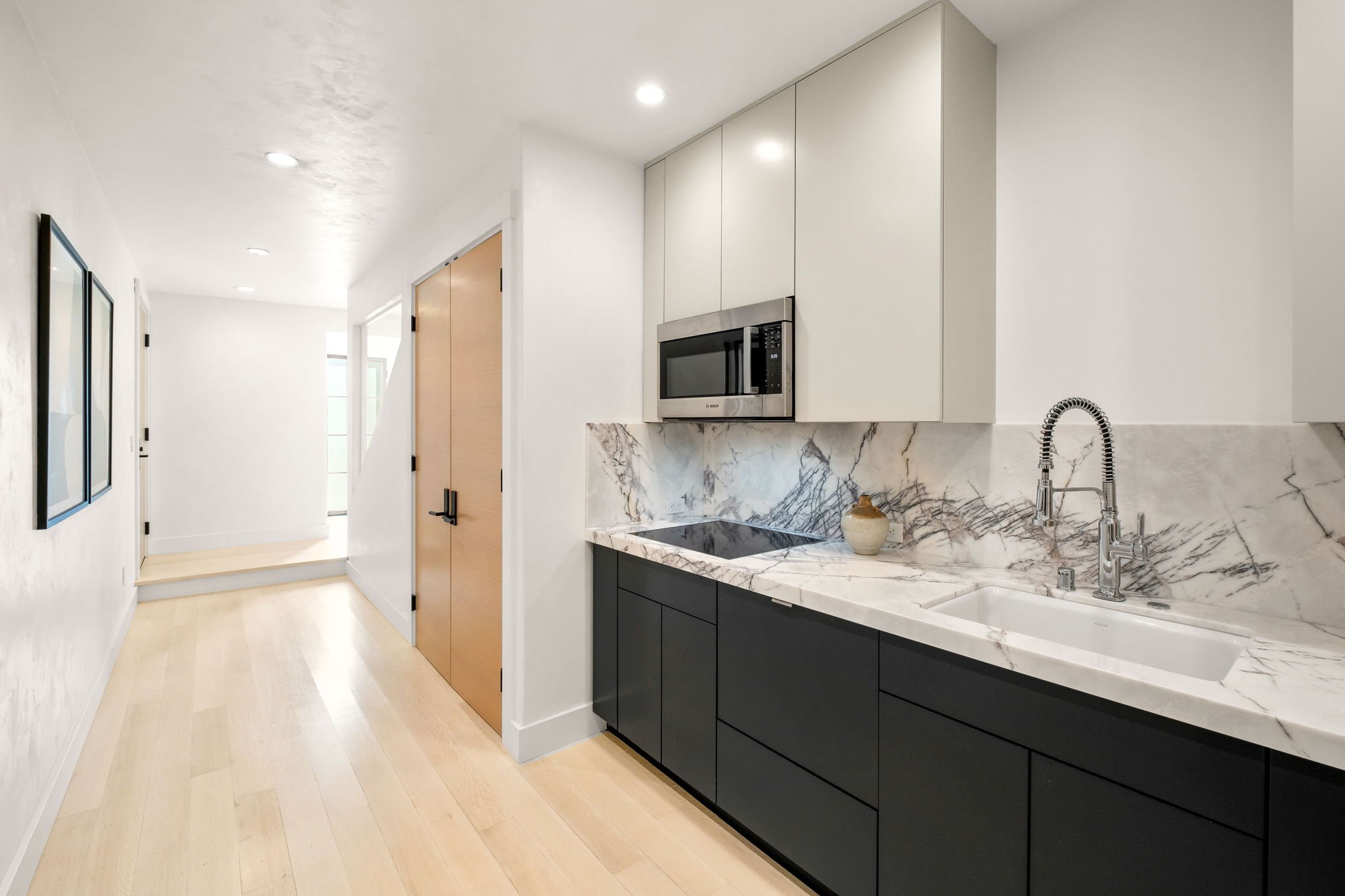 Modern kitchen with white upper cabinets, black lower cabinets, marble backsplash, and countertop, stainless steel microwave and sink with a gooseneck faucet, light wood flooring, and a hallway with framed artwork.