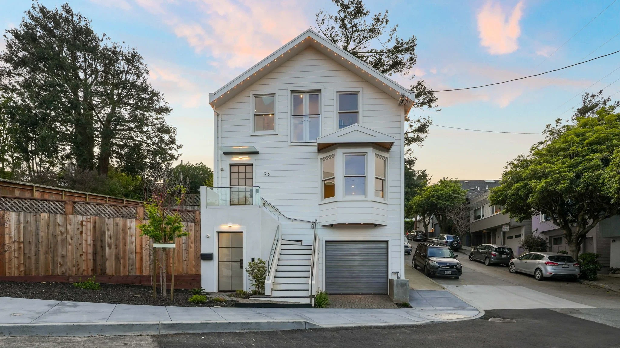 A modern white multi-story house with bay windows, a small front porch, staircase, and a garage, situated on a residential street with parked cars and trees.