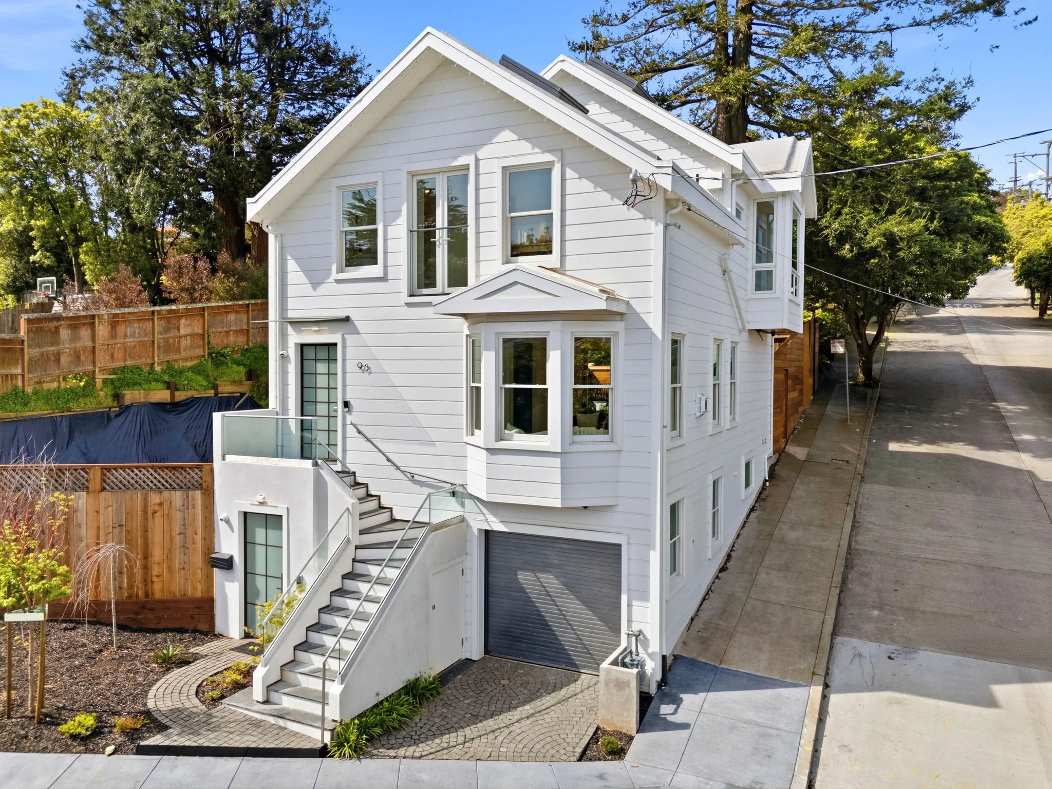 A modern white three-story house with exterior stairs, a garage door, multiple windows, and a small front yard with landscaping, located on a sloped street.